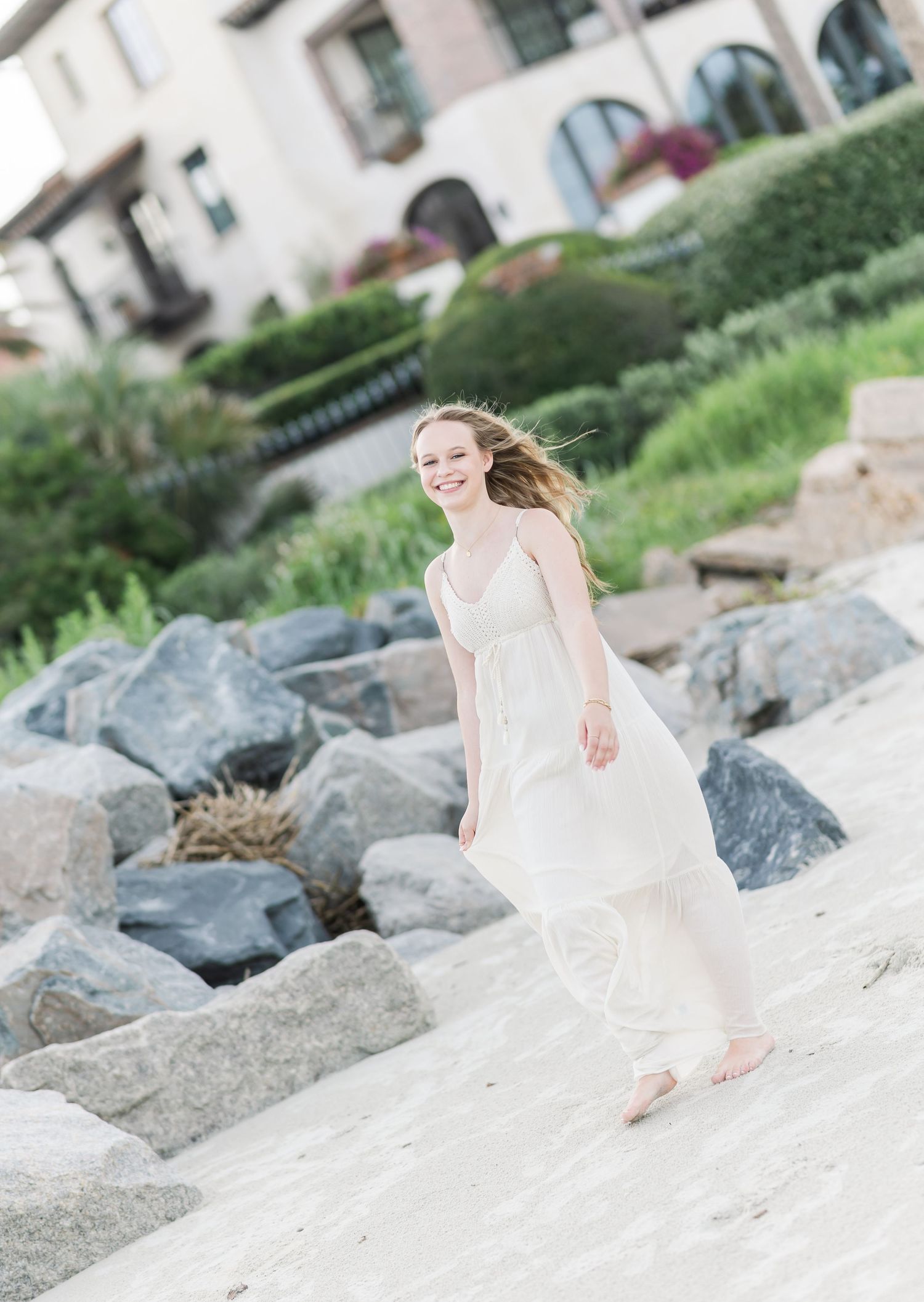 A white dress flows in the wind while walking along rocky beach with Spanish-style architecture behind.