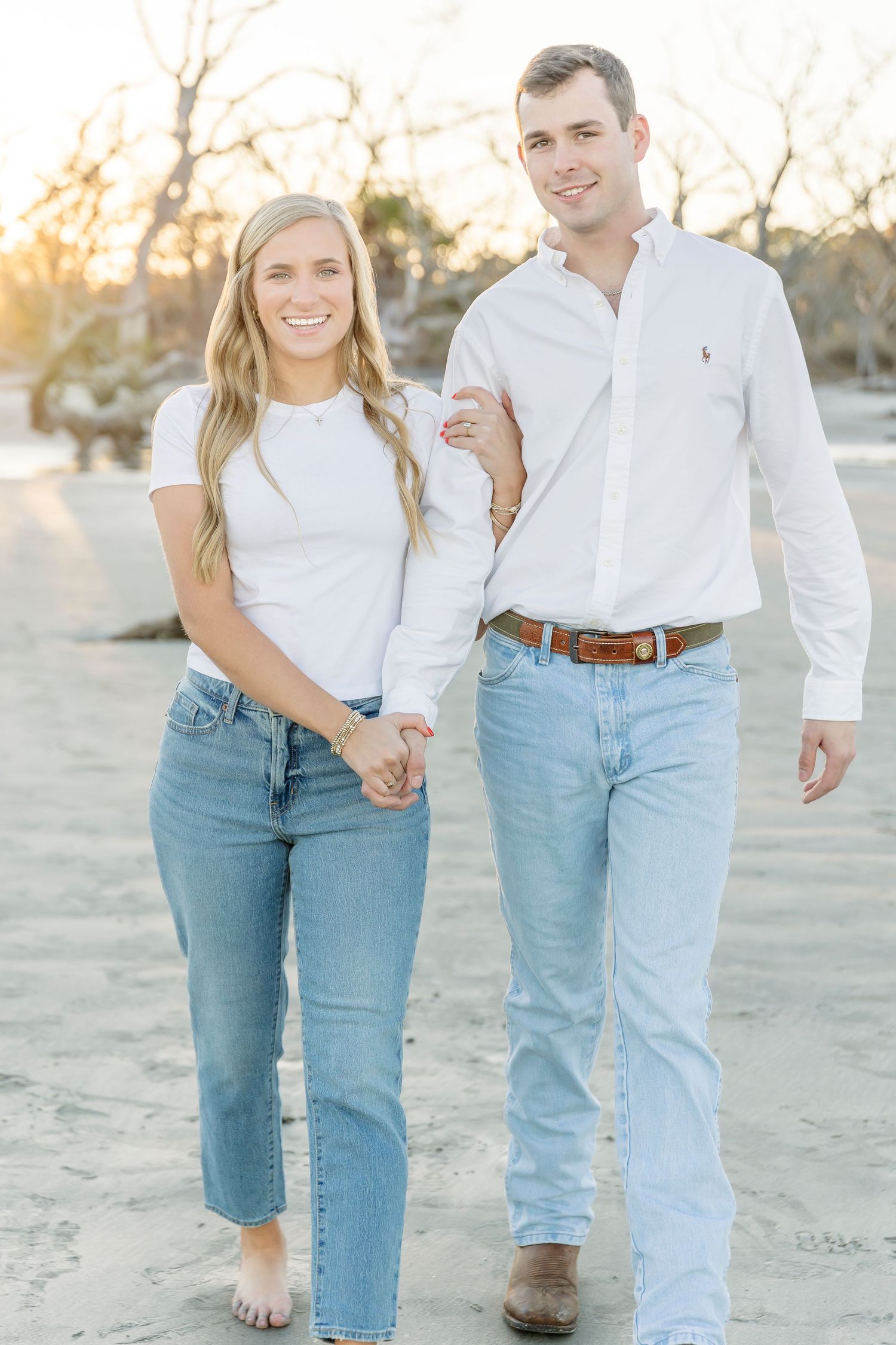 A couple walks barefoot on the beach at sunset wearing matching white tops and blue jeans.