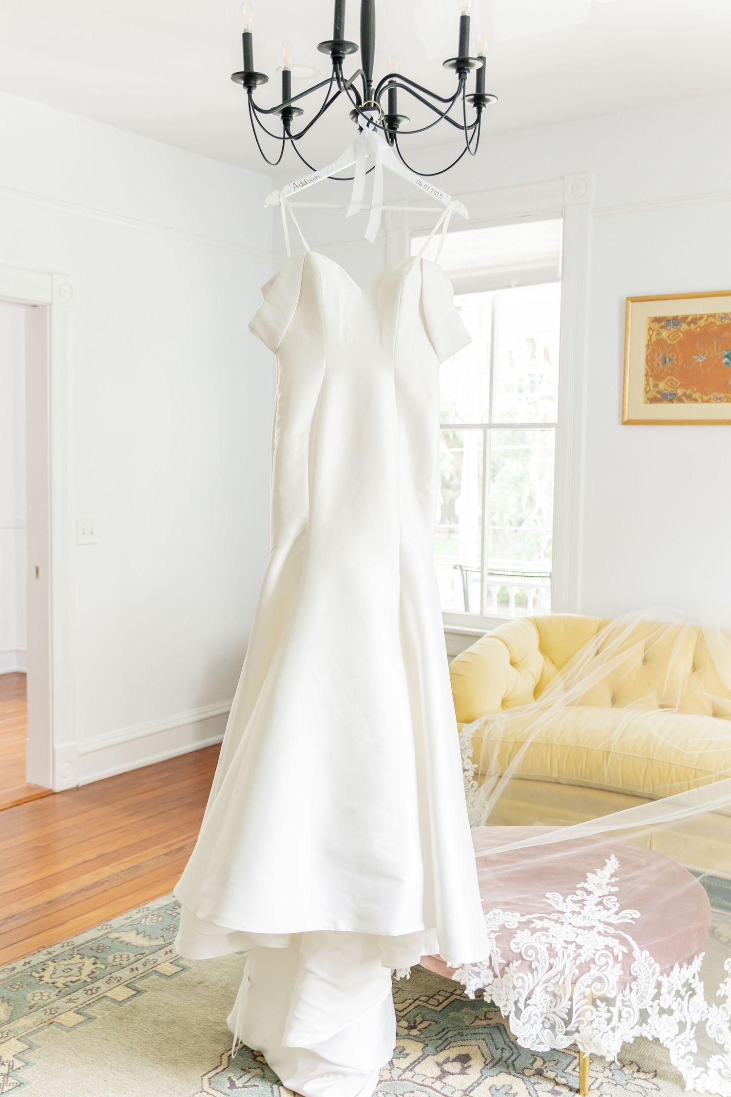 Elegant white wedding dress hanging from chandelier with yellow sofa and window in bright sunlit room.