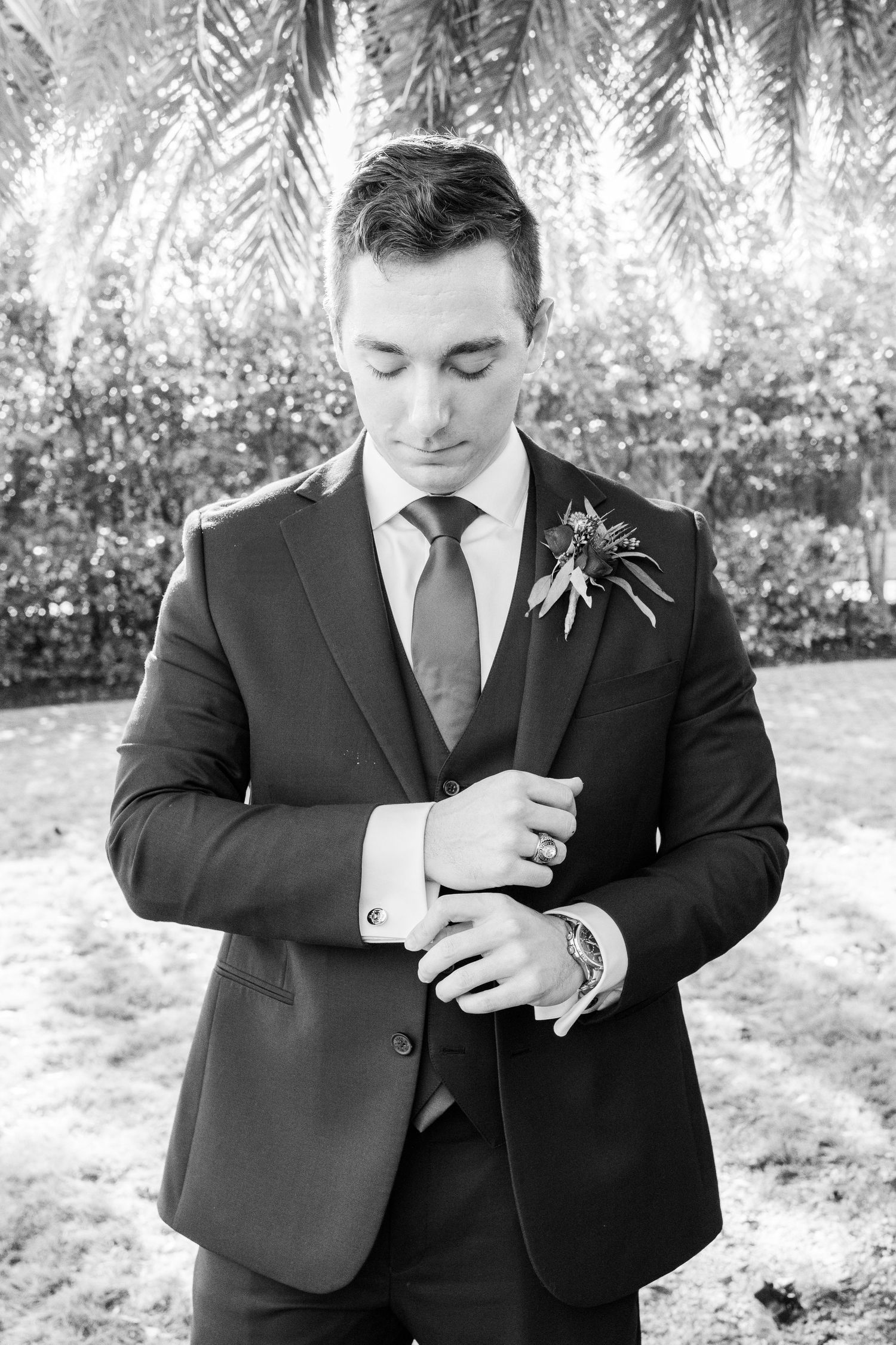 Black and white photo of groom adjusting his suit sleeve under palm trees.