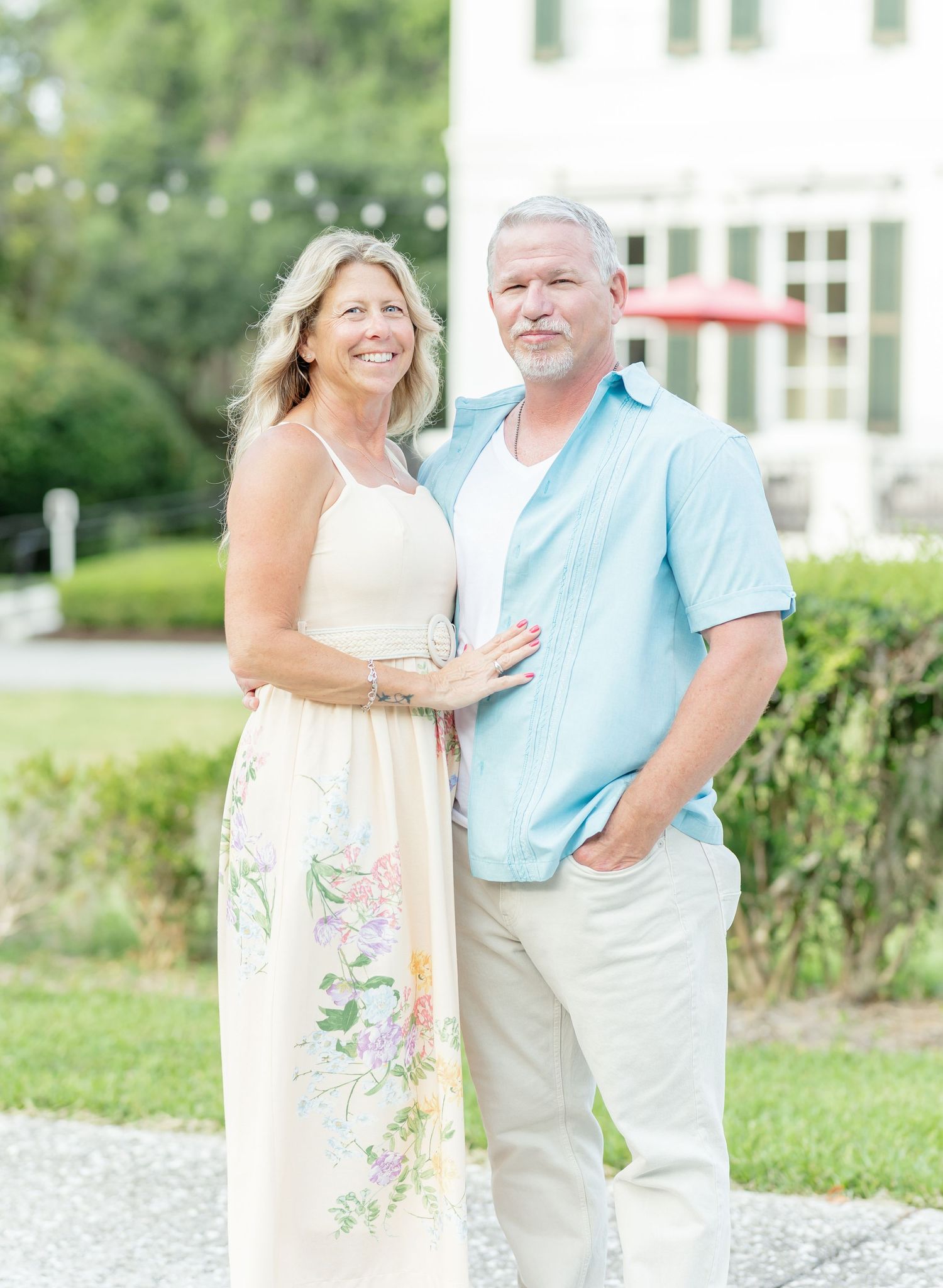 A couple in light summer attire share a tender moment together on a gravel path with greenery in the background.