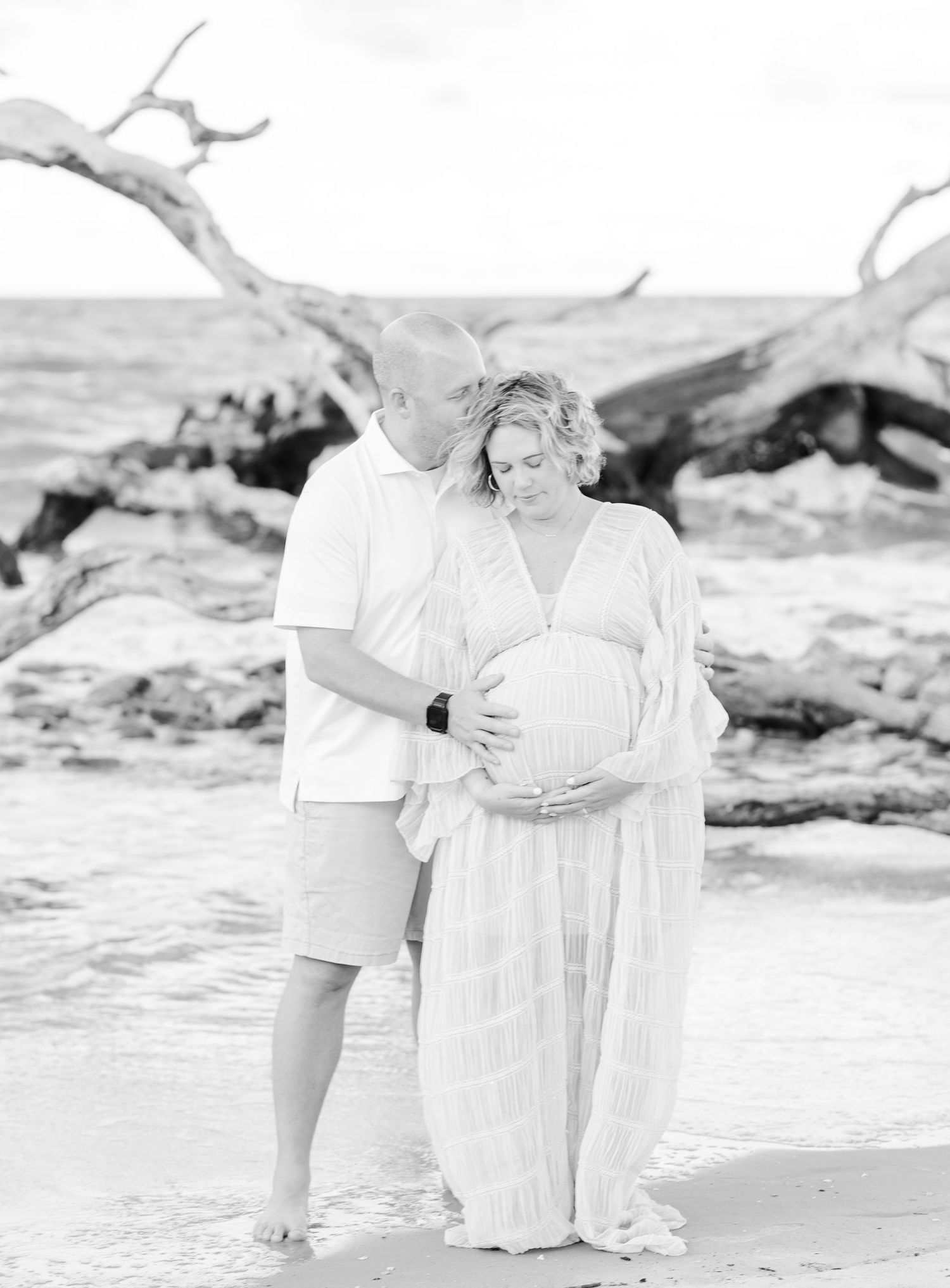 A family dressed in white poses together on a beach with driftwood and waves in the background.