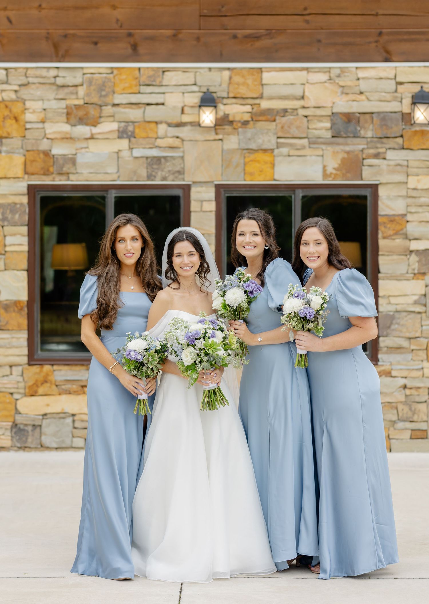 A bridal party in light blue dresses and a white wedding gown pose together against a stone wall exterior.