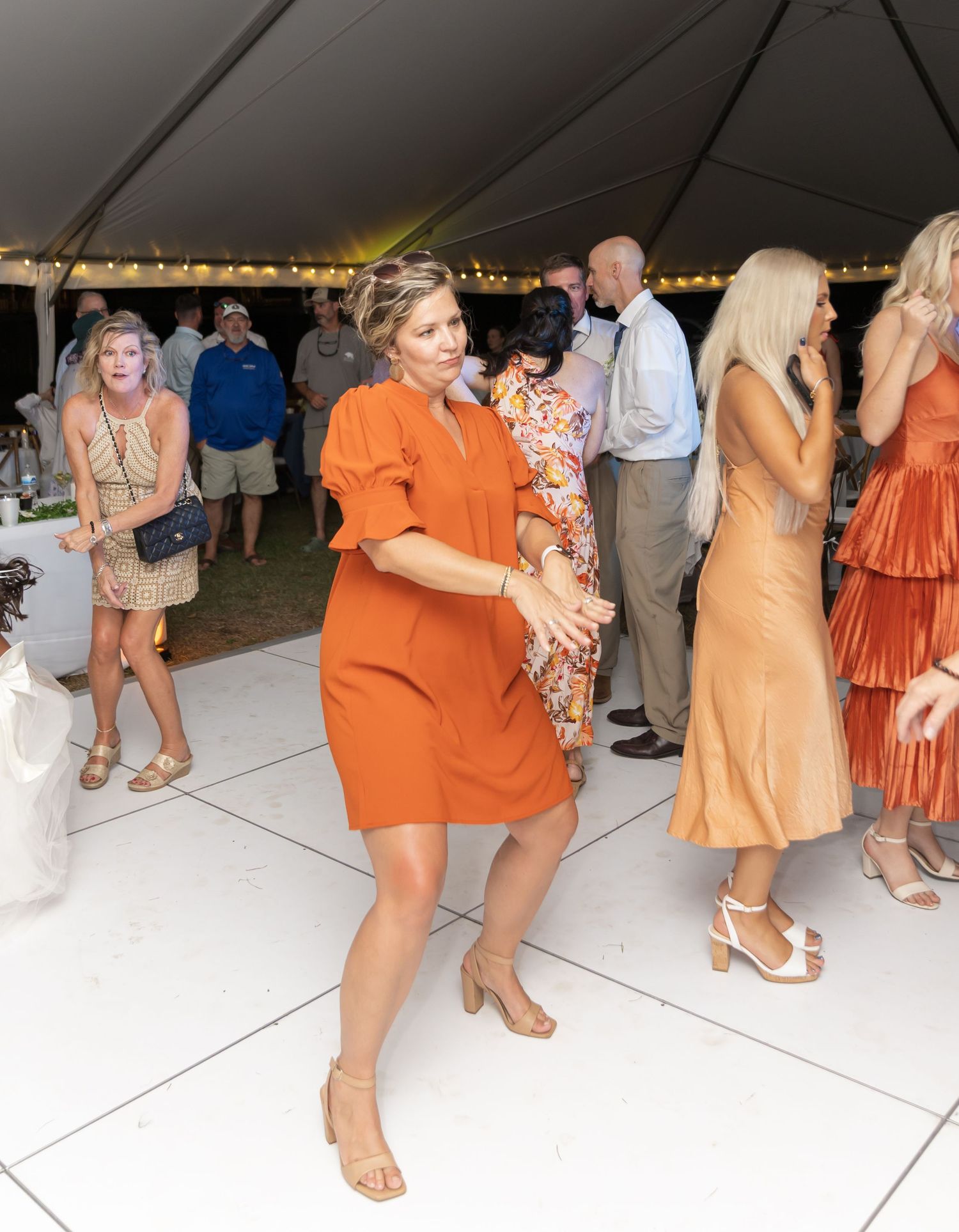 Wedding guests in orange and peach dresses dancing on white dance floor under tent.