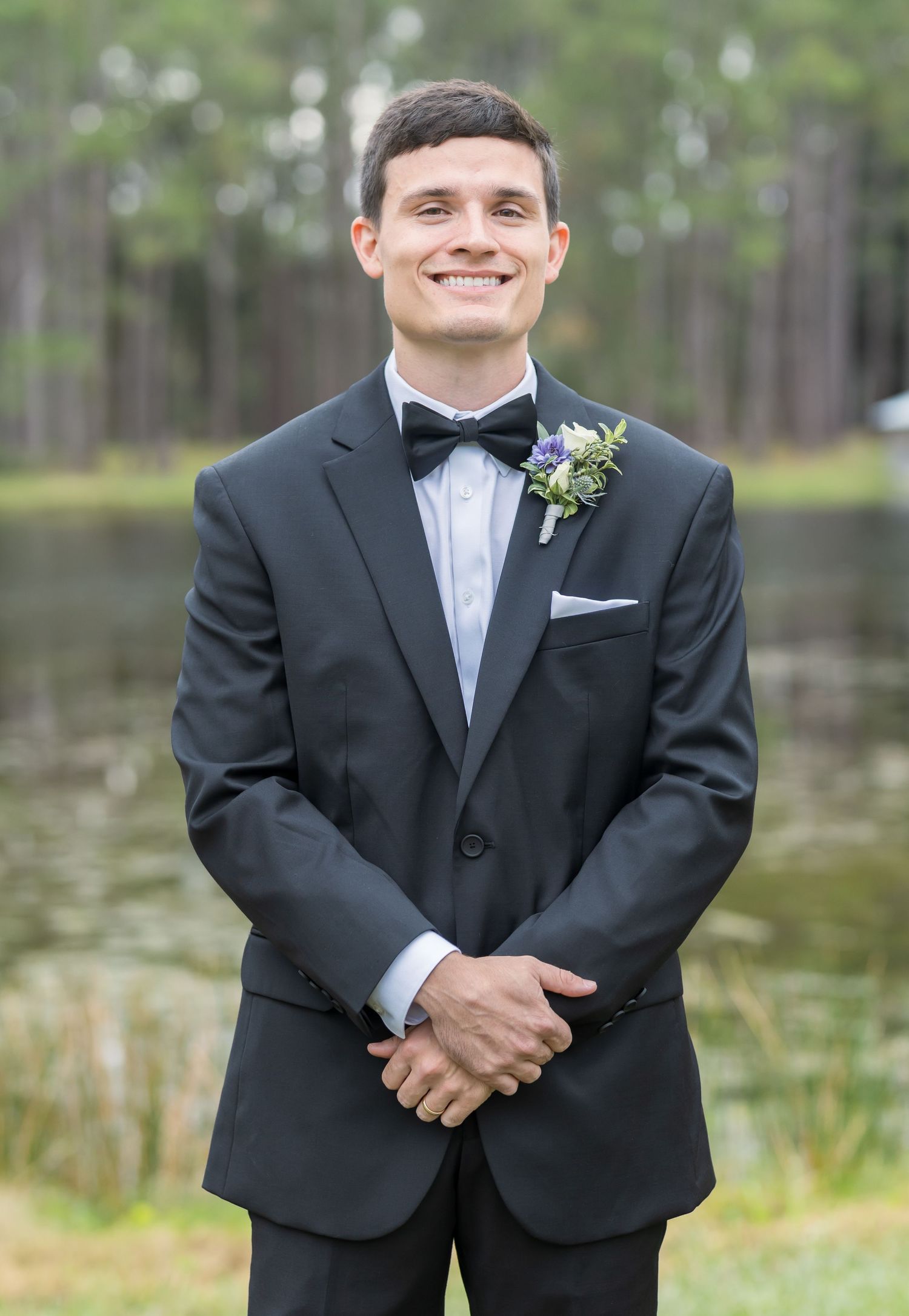 Groom in classic black tuxedo and bowtie poses outdoors against a wooded lake background.