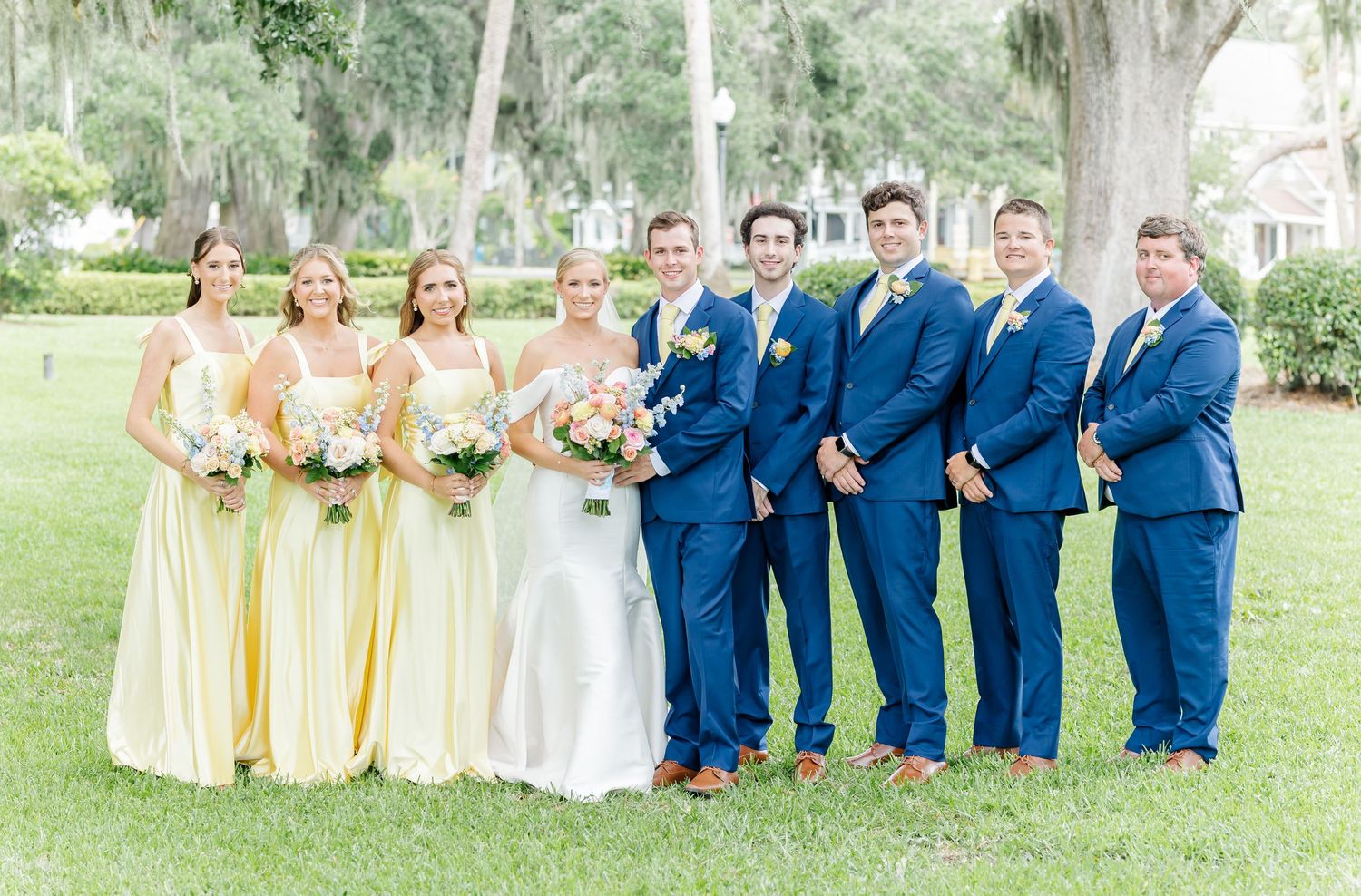 Wedding party in navy blue suits and yellow dresses poses together on grassy lawn for formal group photos.