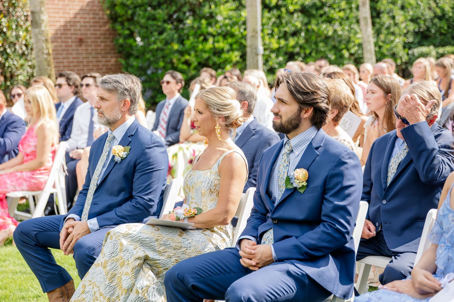 Wedding guests in navy suits and summer dresses sit outdoors during a garden ceremony on a sunny day.