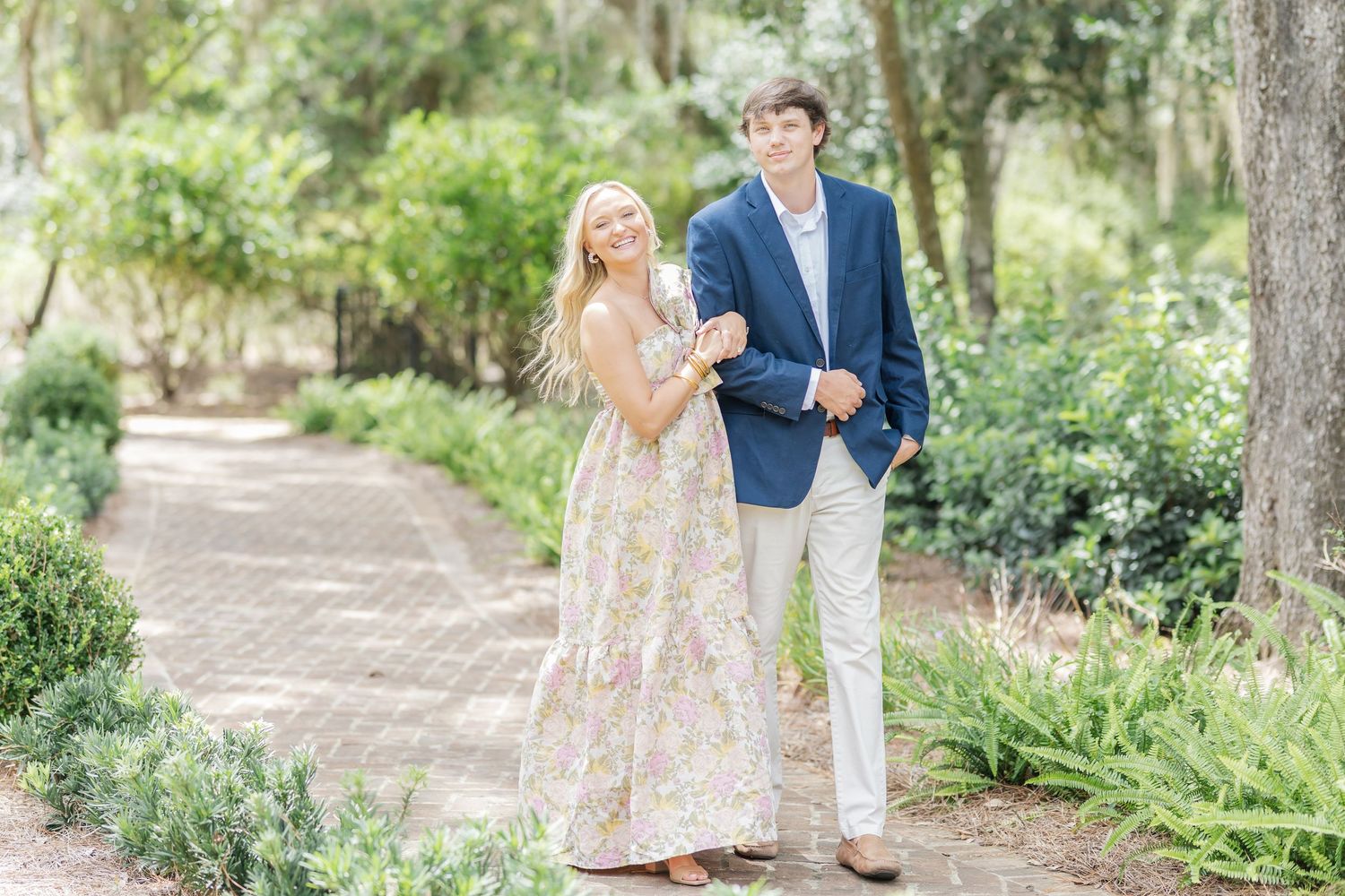 A couple poses together on a brick pathway in a southern garden with hanging moss overhead.