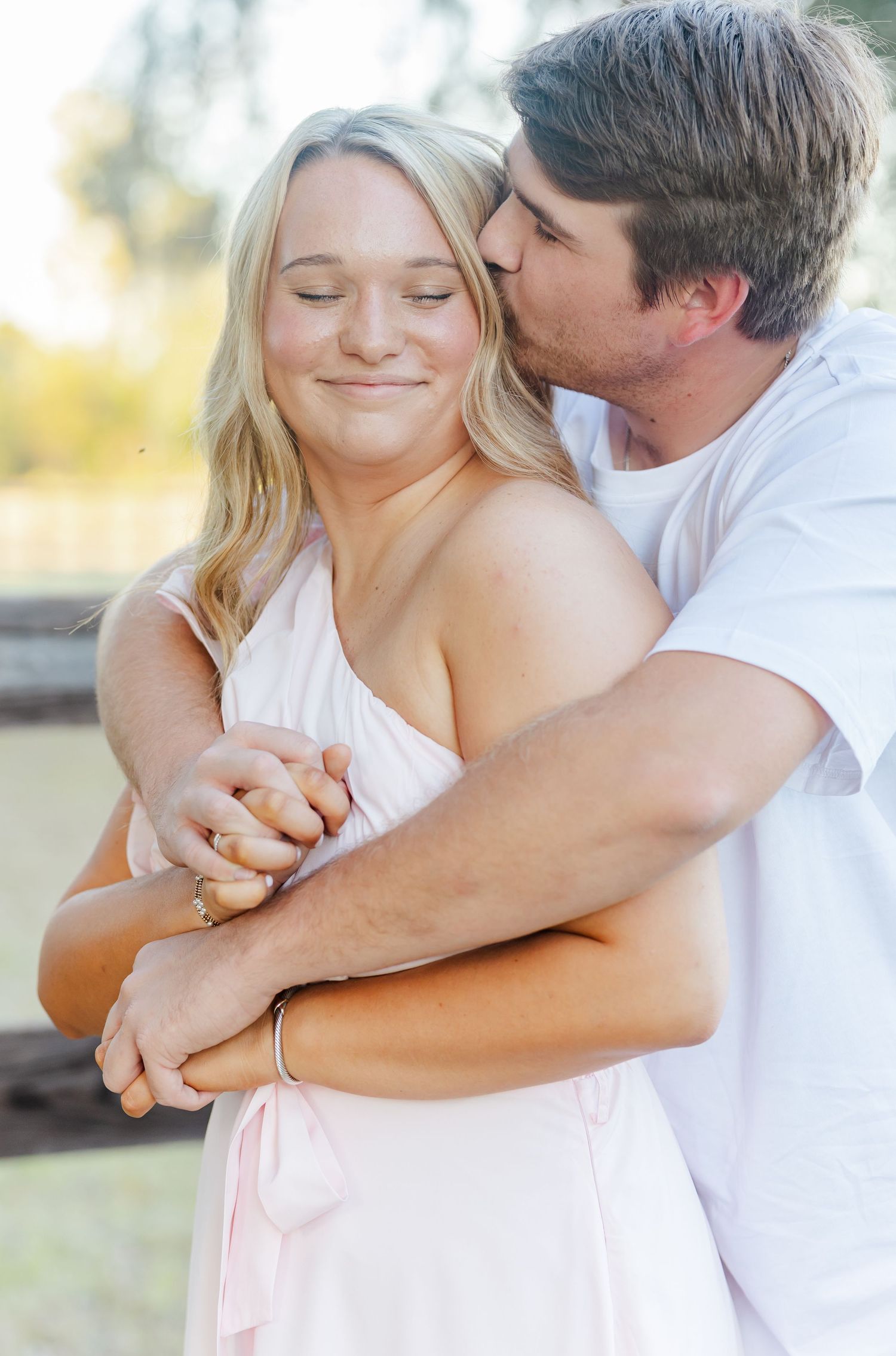 A romantic couple embraces tenderly in soft natural lighting wearing white and pink clothing.