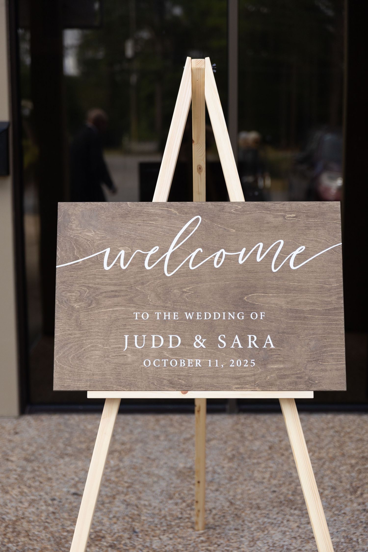 Rustic wooden wedding welcome sign with white calligraphy displayed on white easel.