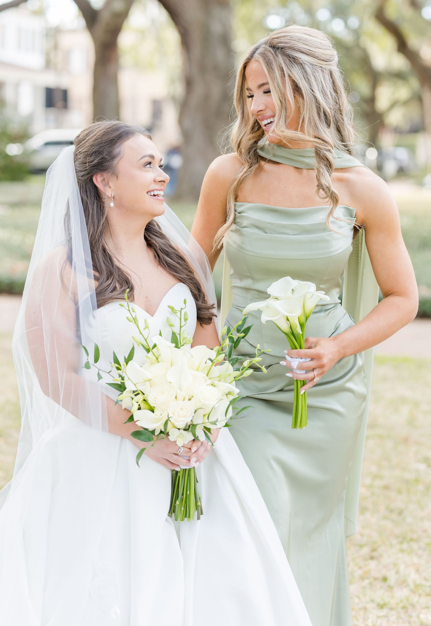Smiling bride in white gown with veil and bridesmaid in sage green dress share a joyful moment outdoors.