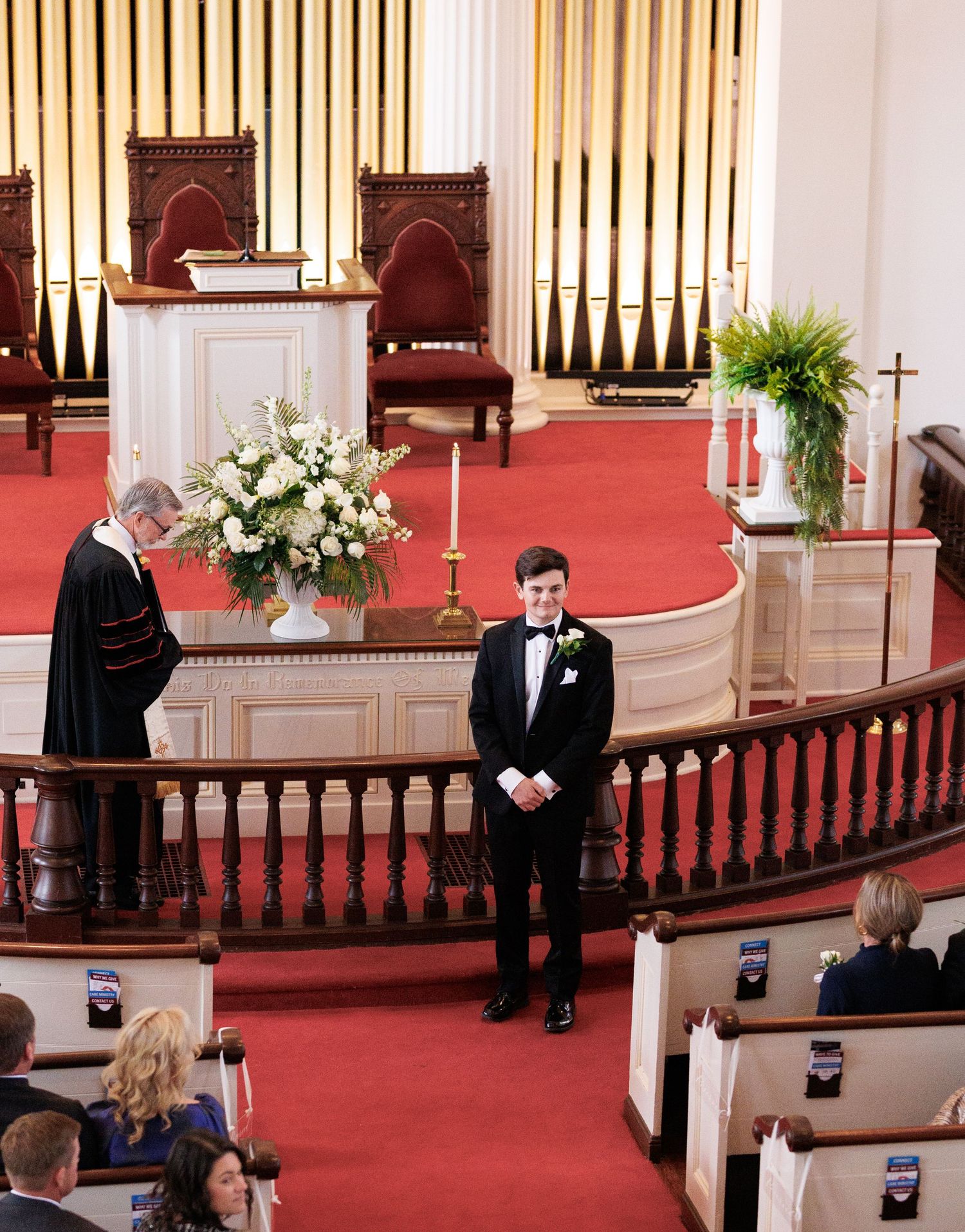 Groom in tuxedo stands at church altar awaiting bride, with officiant and red carpet aisle during wedding ceremony.