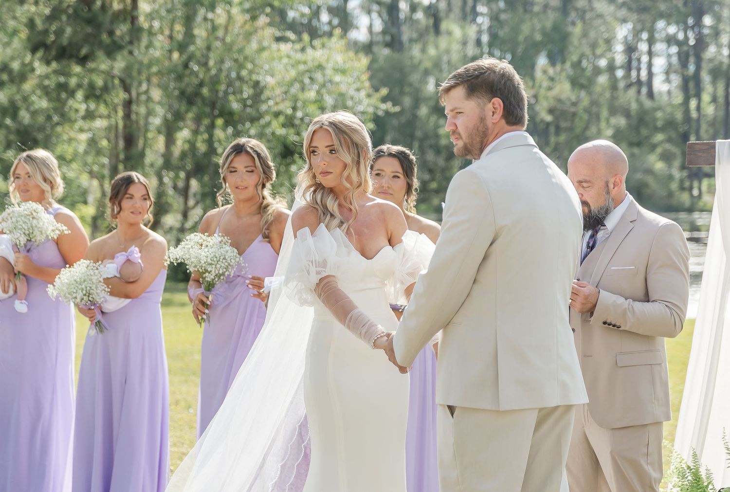 An outdoor spring wedding ceremony with bridesmaids in lavender dresses and light colored decor.