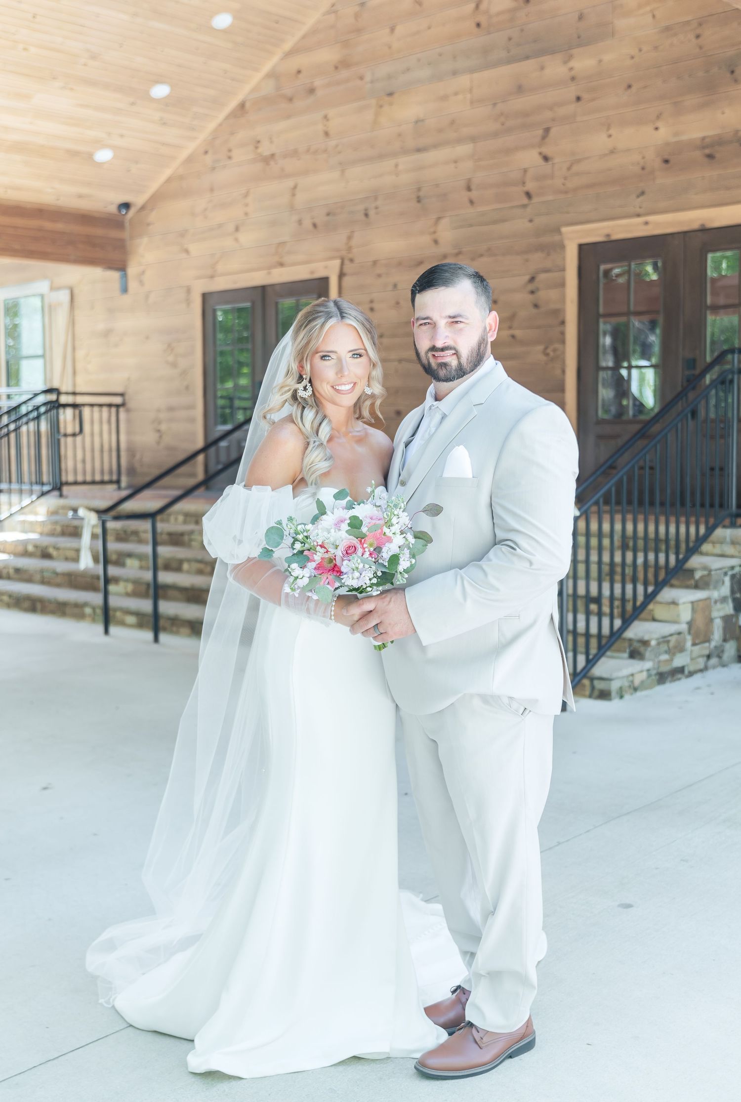 Elegant wedding couple poses on porch of wooden building with pink and white floral bouquet.
