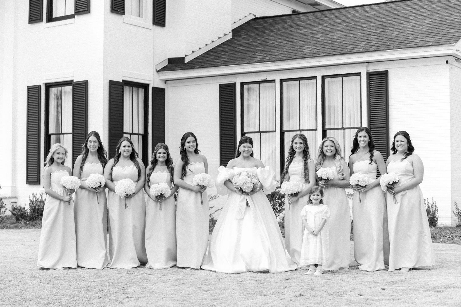 Black and white photo of bride in ball gown with bridesmaids in champagne dresses and flower girl outside white venue.