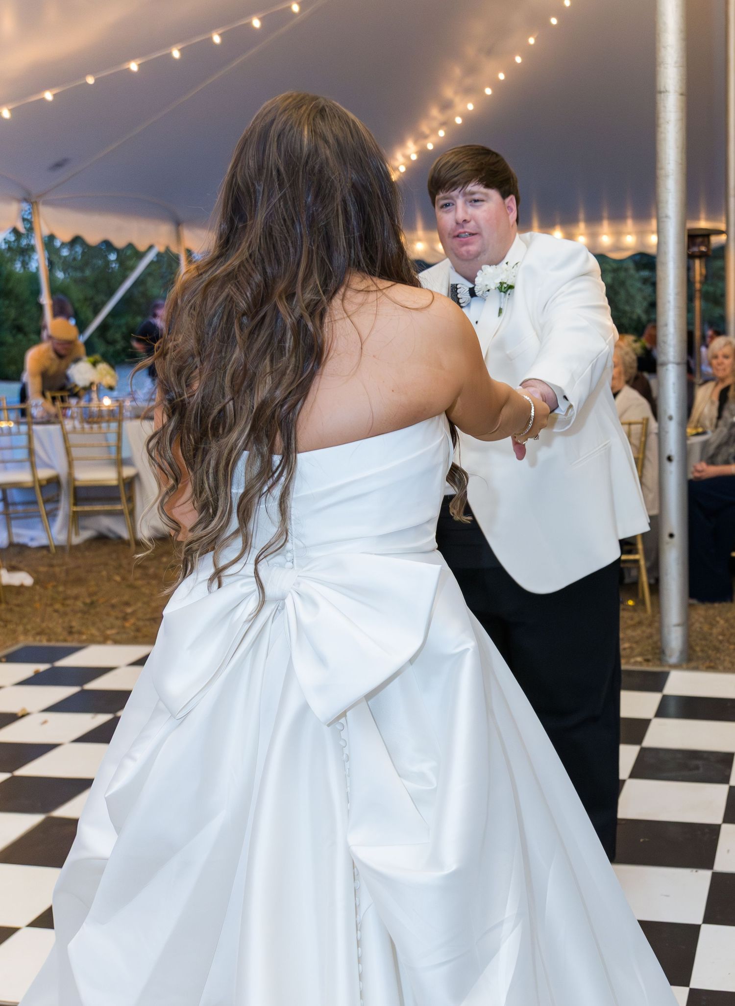 Bride in strapless gown with large bow dancing with groom in white jacket on checkered floor.