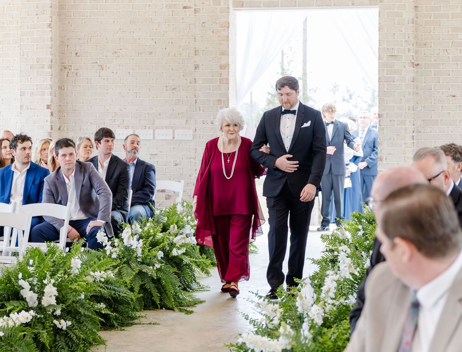 Elderly woman in red escorted down floral-lined wedding aisle by young man in tuxedo.