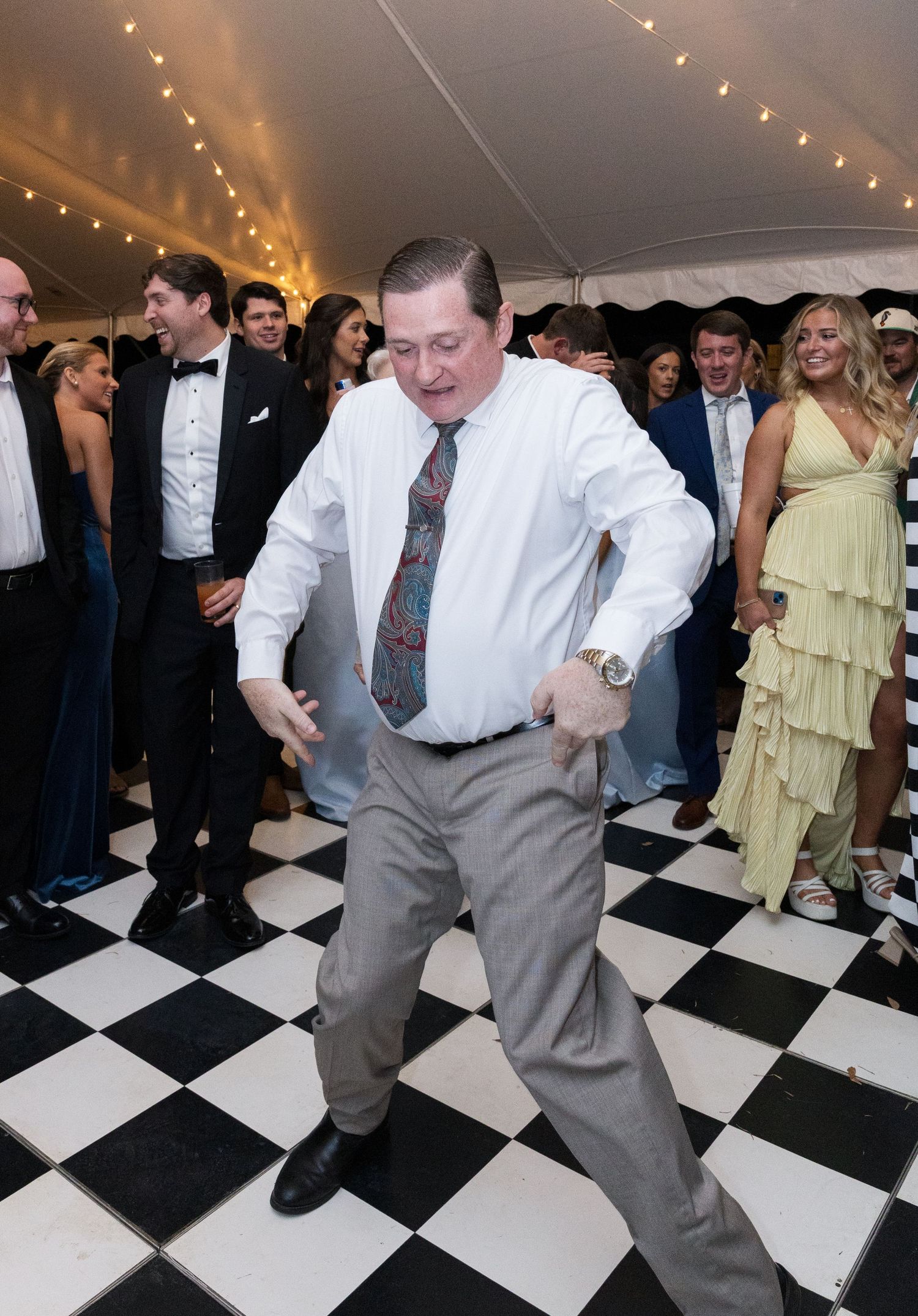 Enthusiastic wedding guest shows off dance moves on a checkered dance floor surrounded by cheering guests.