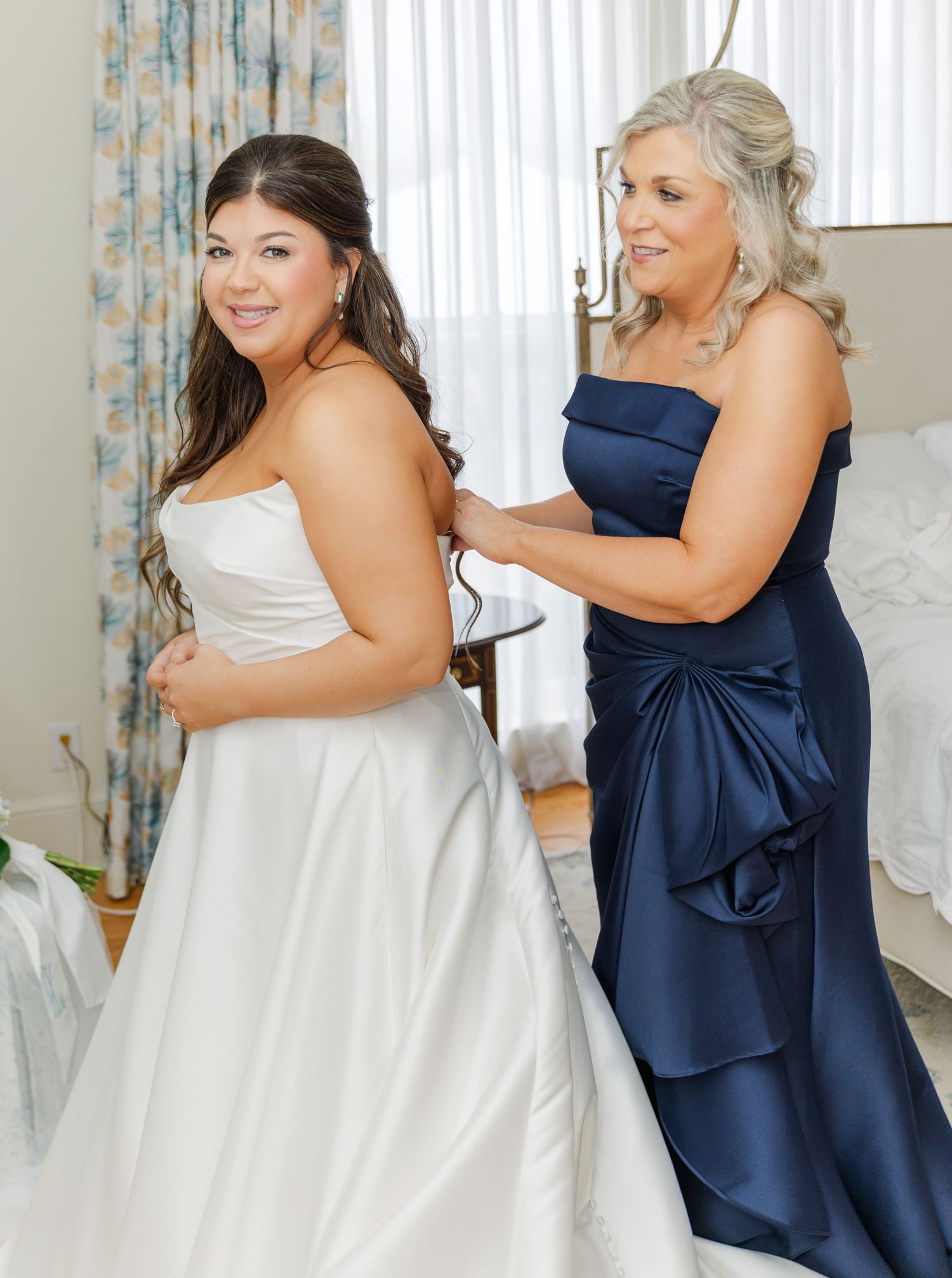 Bride in strapless white gown gets dressed with help from woman in navy blue strapless dress.