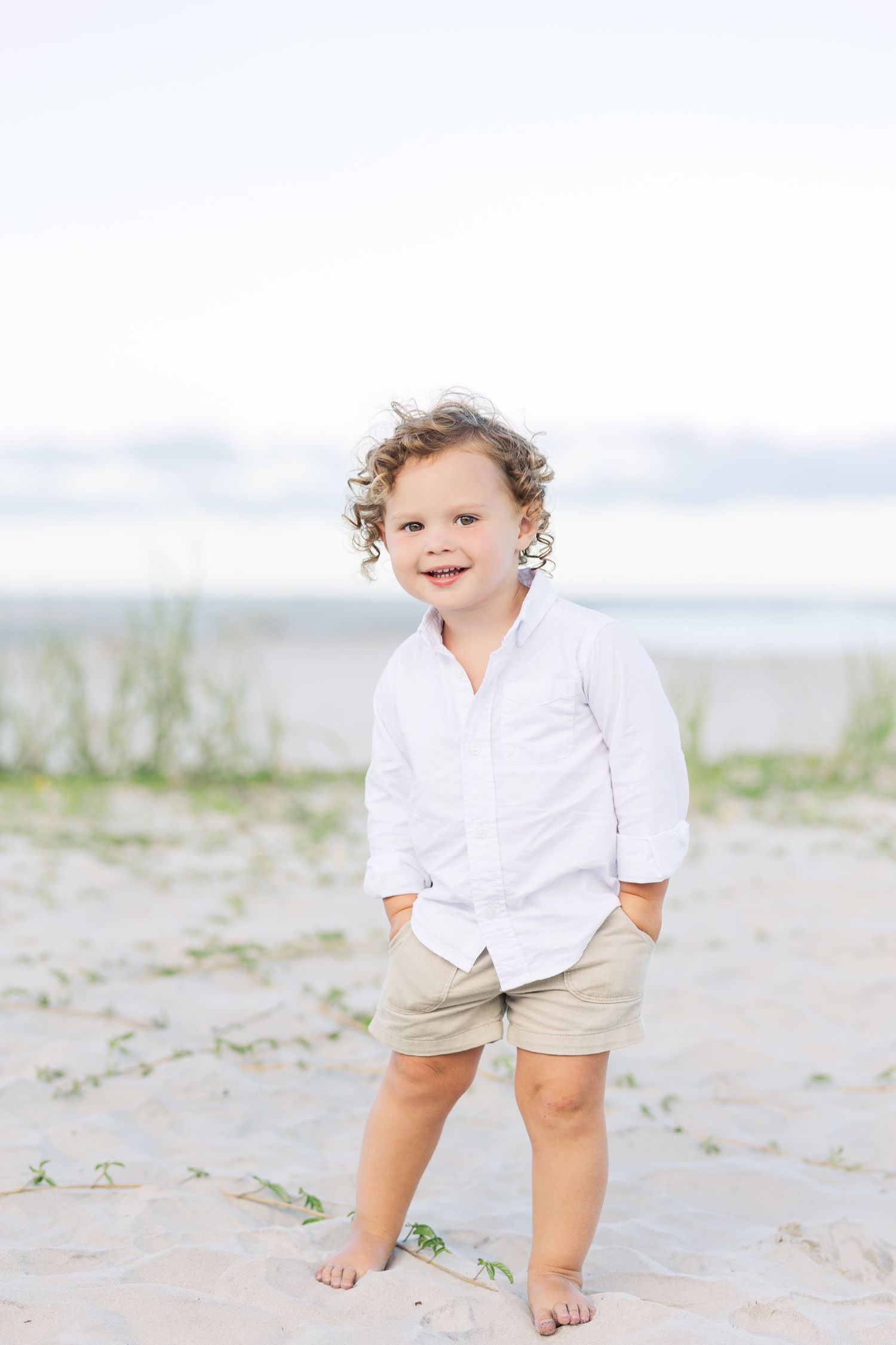 A small child in a white button-down shirt and khaki shorts stands on a sandy beach.