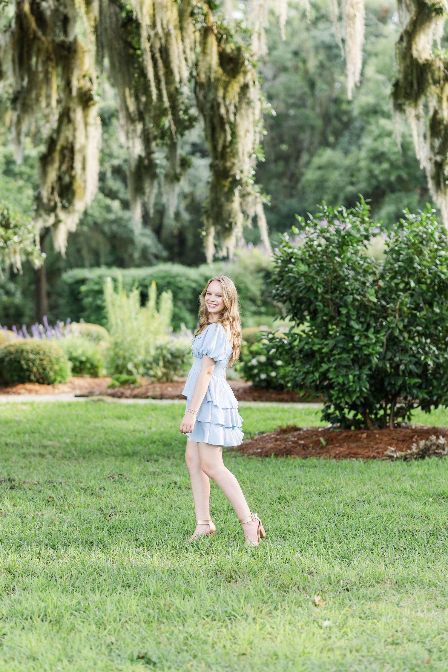 A light blue dress catches the breeze in a garden setting with Spanish moss draped trees.