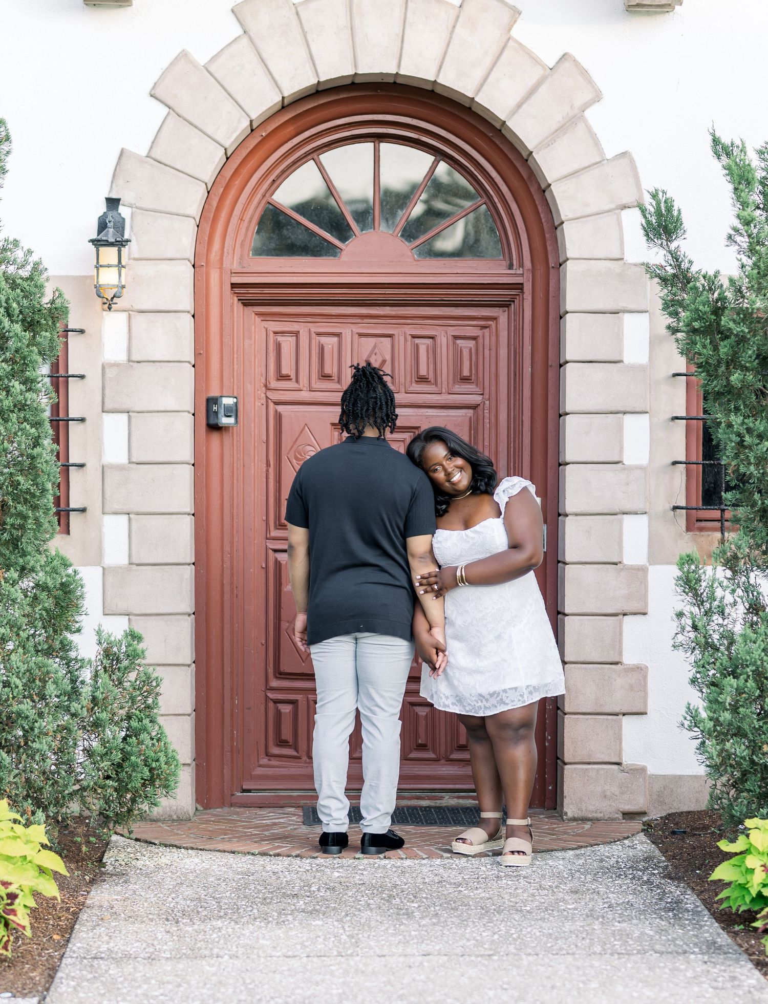 A couple stands hand in hand before a grand arched red door, the woman in a white dress leaning affectionately on her partner.