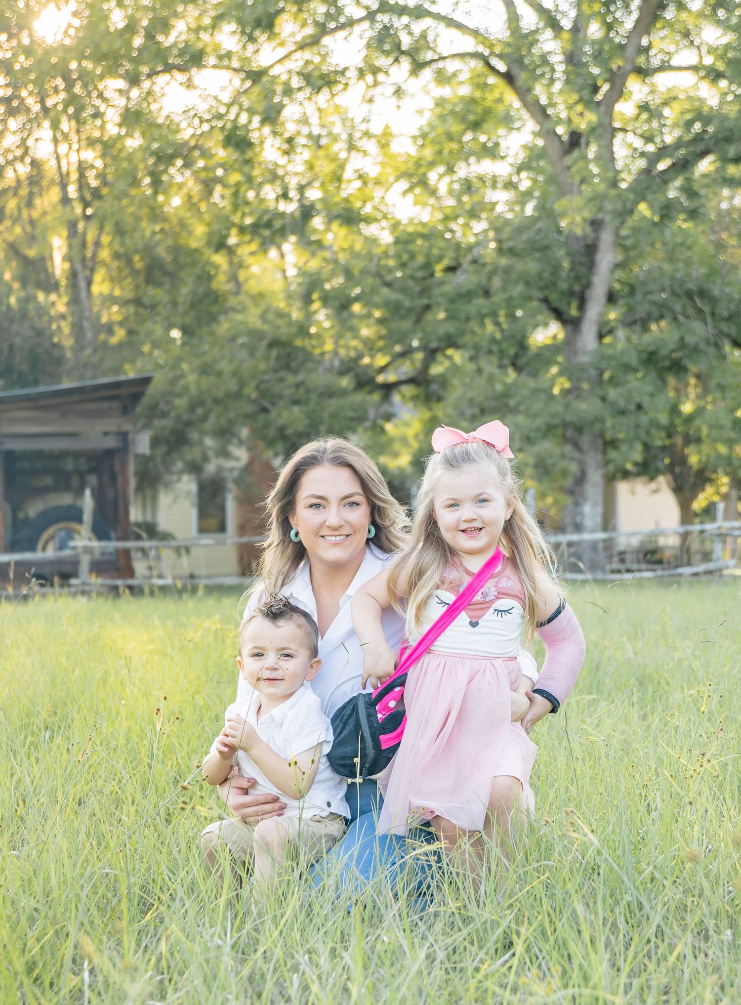 Family sits together in a sunny meadow with tall grass and trees during golden hour in casual outdoor portrait setting.