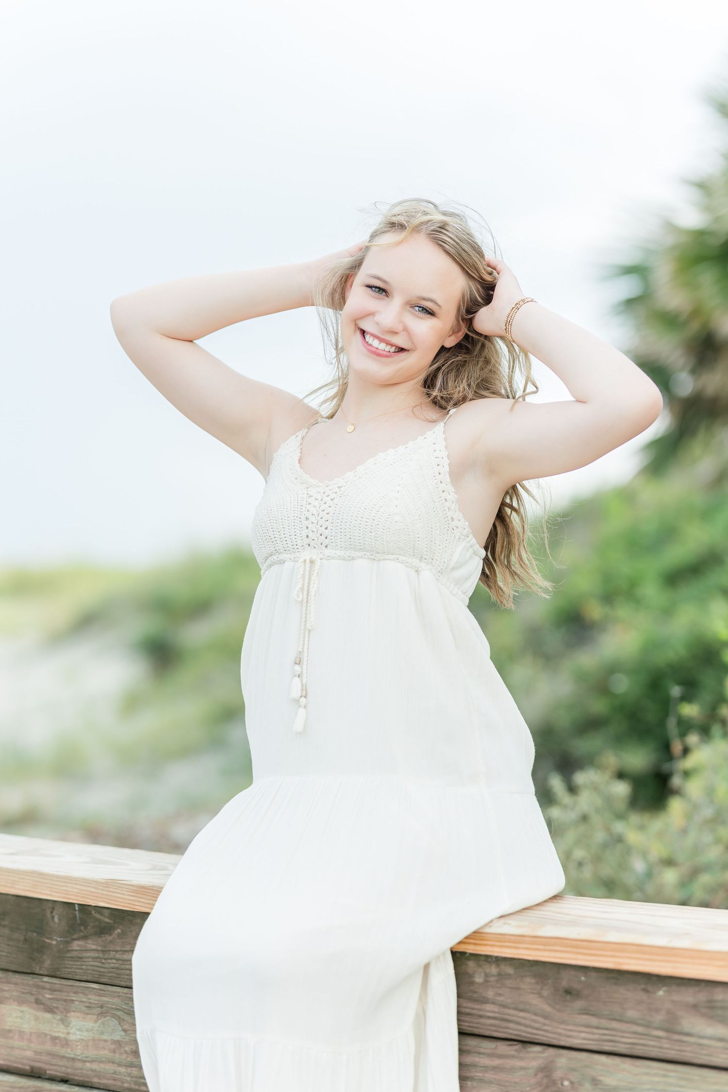Person in white sundress poses playfully on wooden railing outdoors with greenery in background.