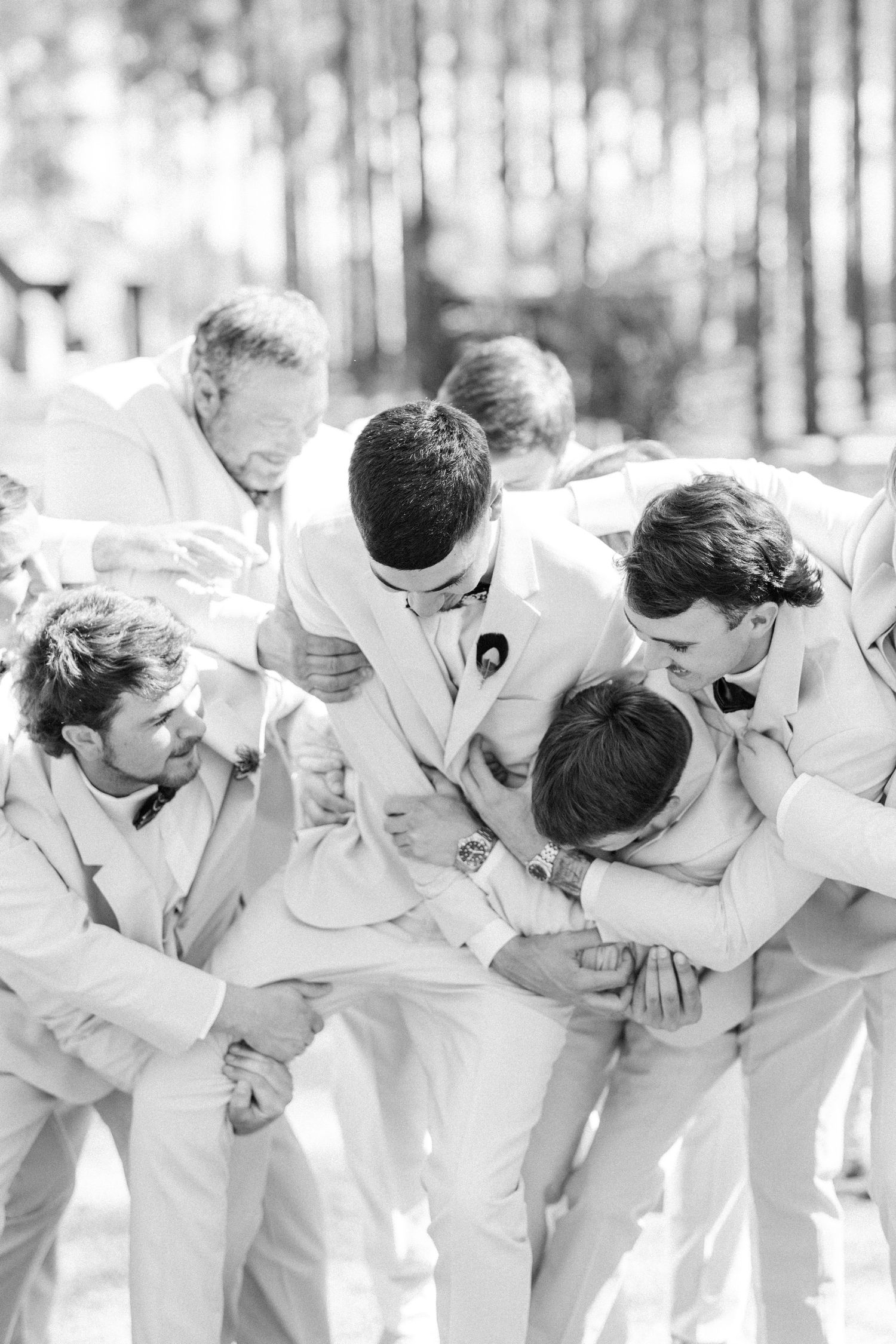 Black and white photo of groom being playfully tackled by groomsmen in matching light suits.