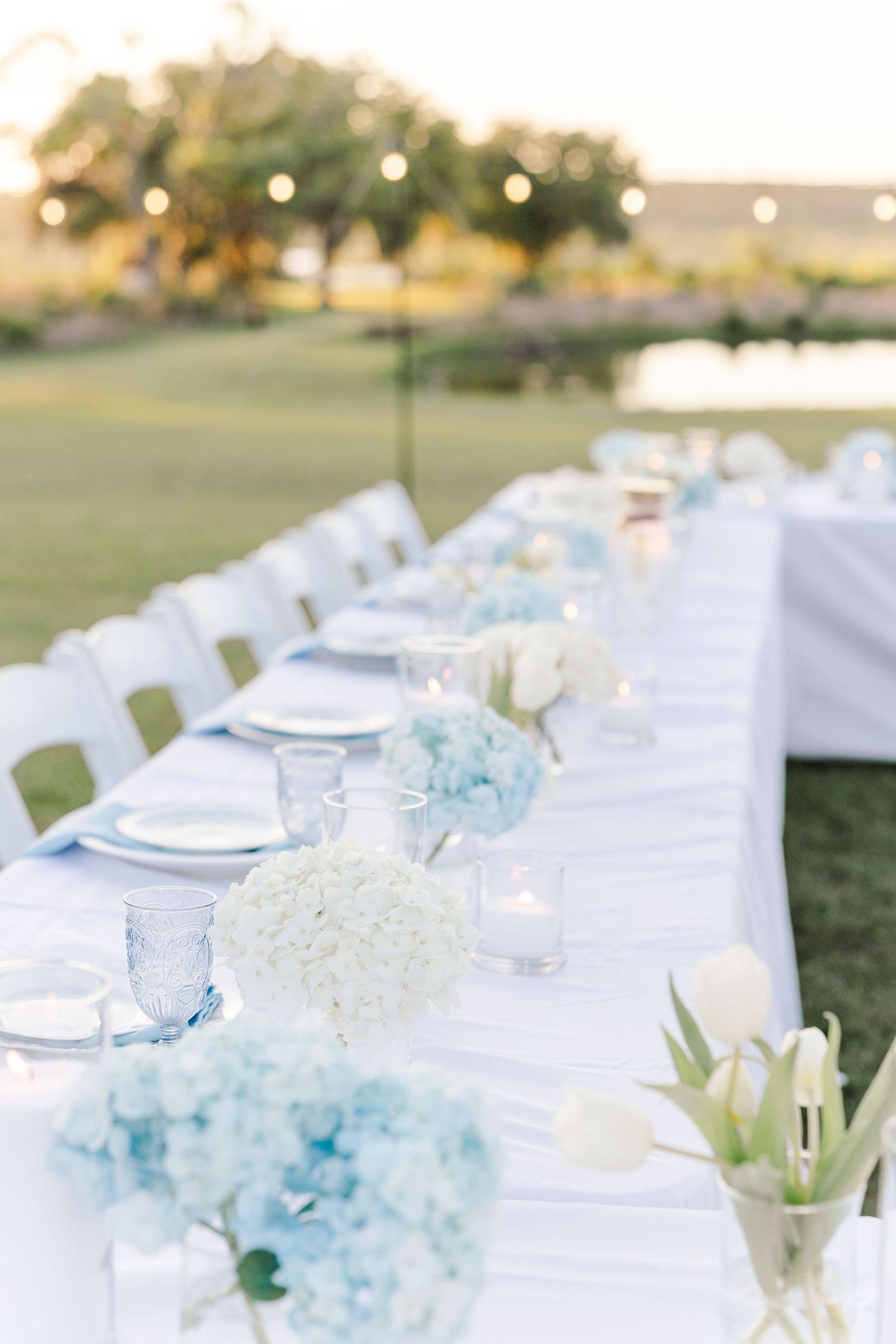 Elegant outdoor wedding tablescape with blue hydrangeas, white tulips, candles, and blue glassware.