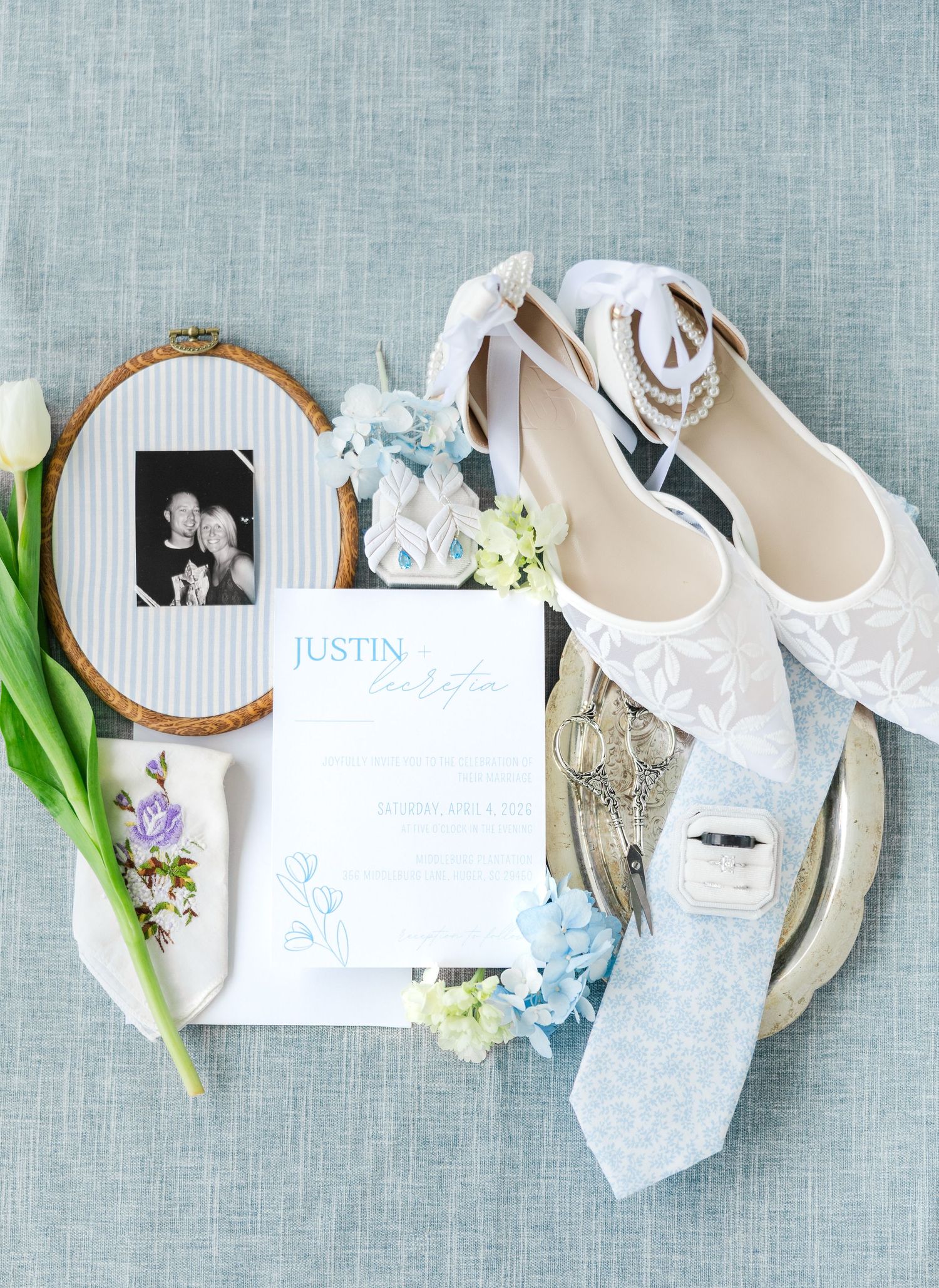 Elegant wedding flat lay with lace shoes, invitation, blue tie, rings, and florals.