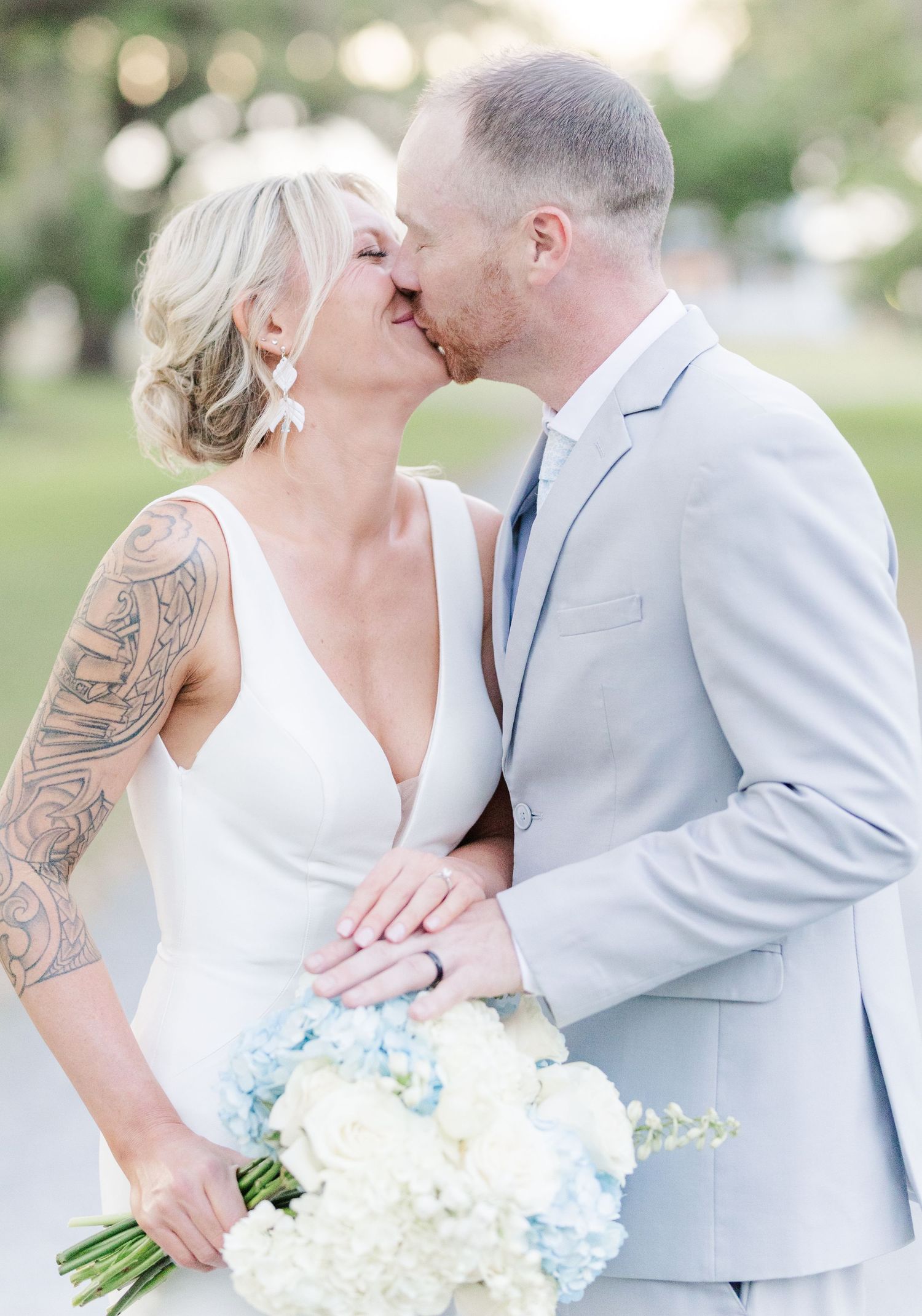 Blonde bride with arm tattoo kissing groom in light gray suit holding blue and white bouquet outdoors.