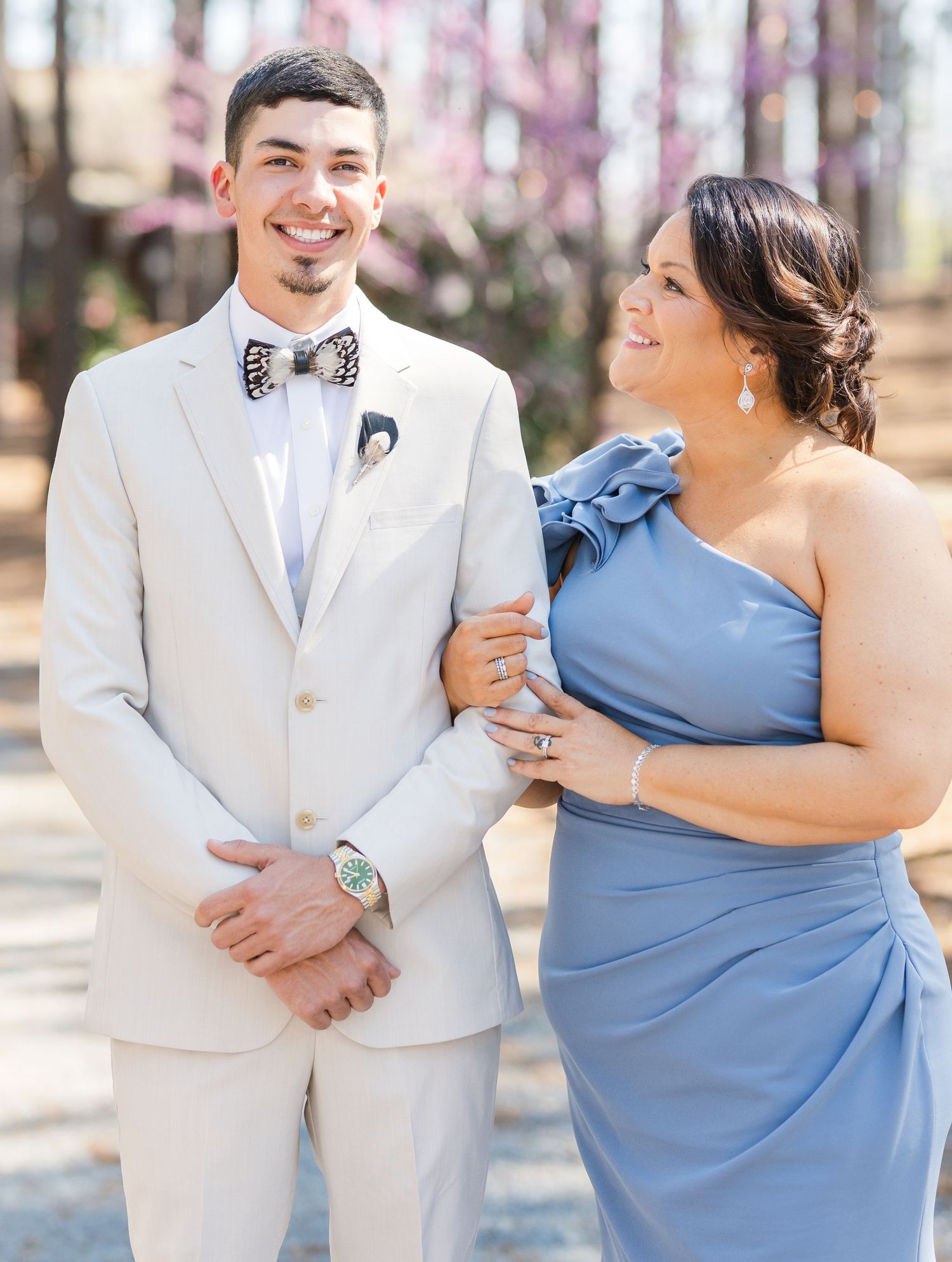 Smiling groom in tan suit with bow tie poses with woman in one-shoulder blue gown at outdoor wedding.
