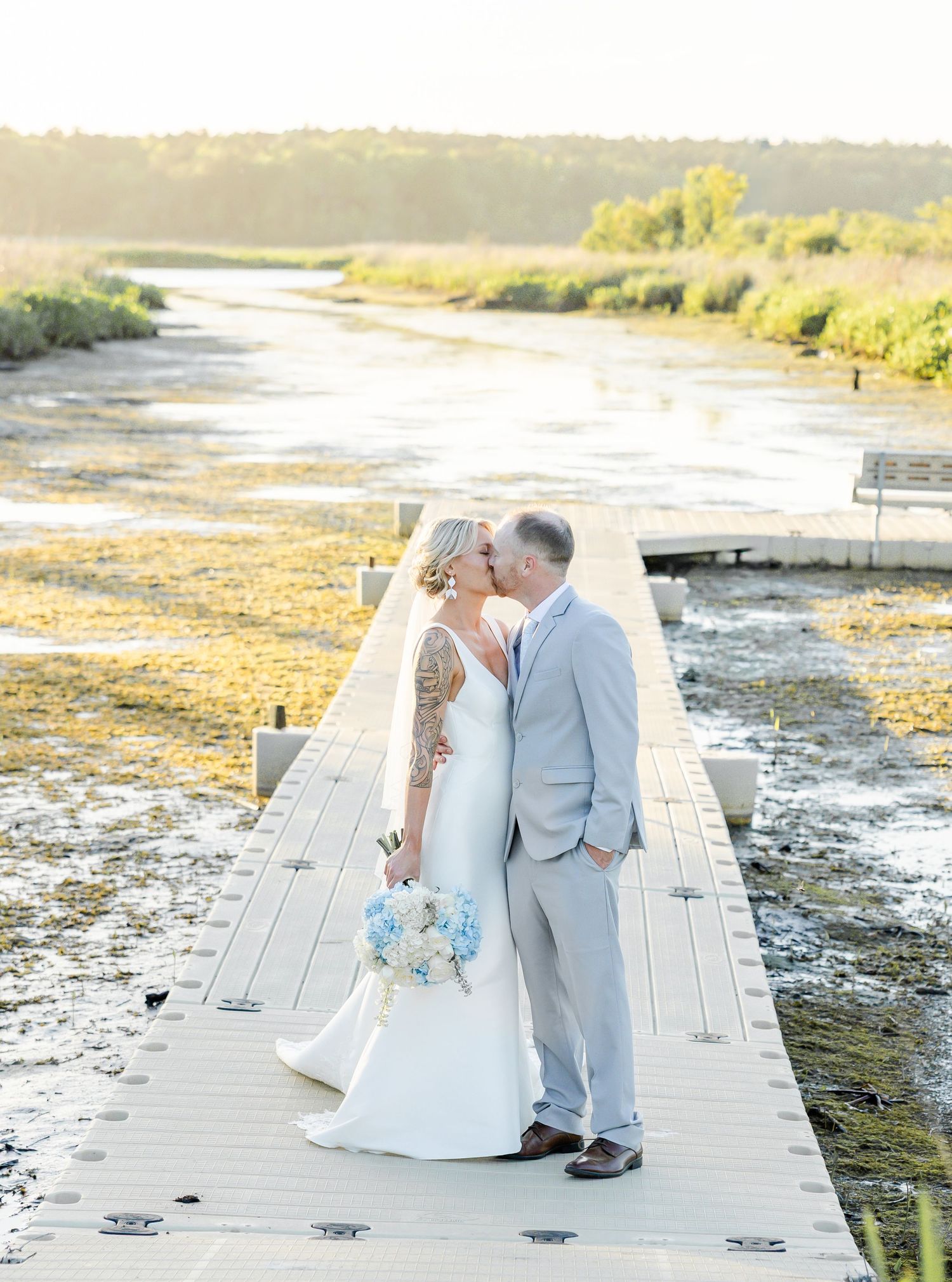 Bride and groom sharing a kiss on a wooden dock overlooking a scenic marsh at sunset.