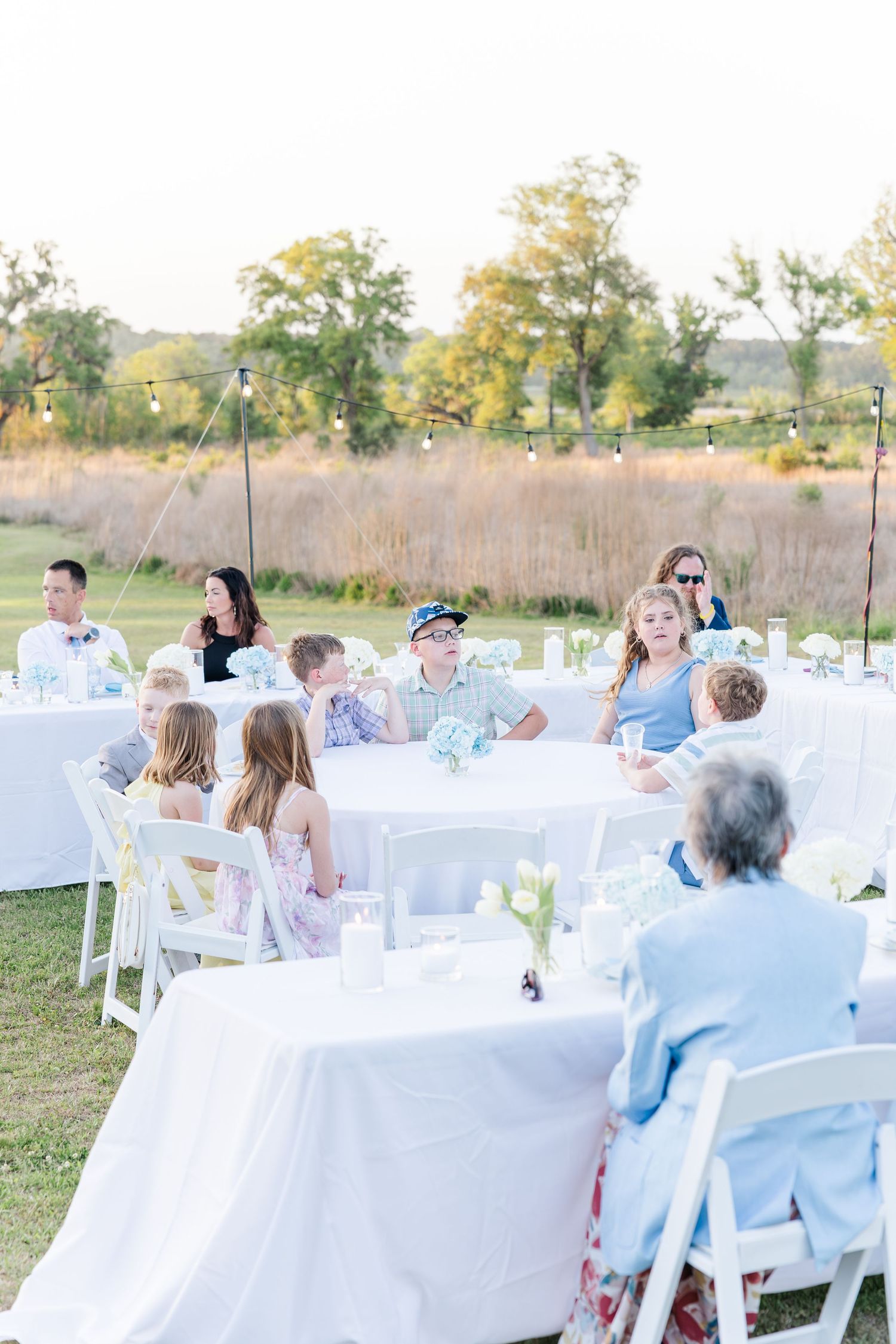 Outdoor wedding reception with guests seated at white-draped round tables under string lights at dusk.