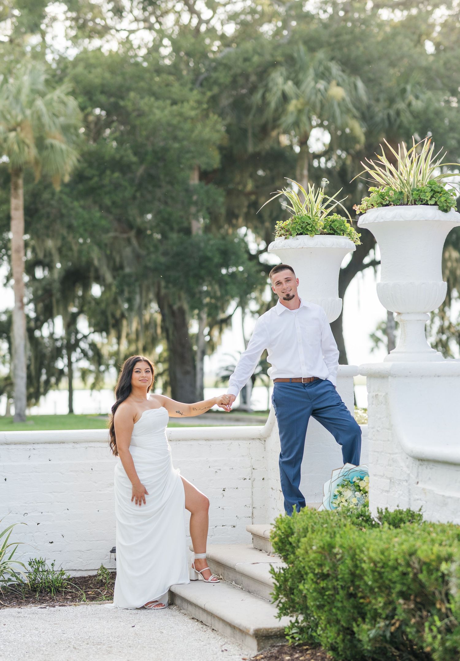 Engaged couple holding hands by ornate white fountain surrounded by palm trees outdoors.