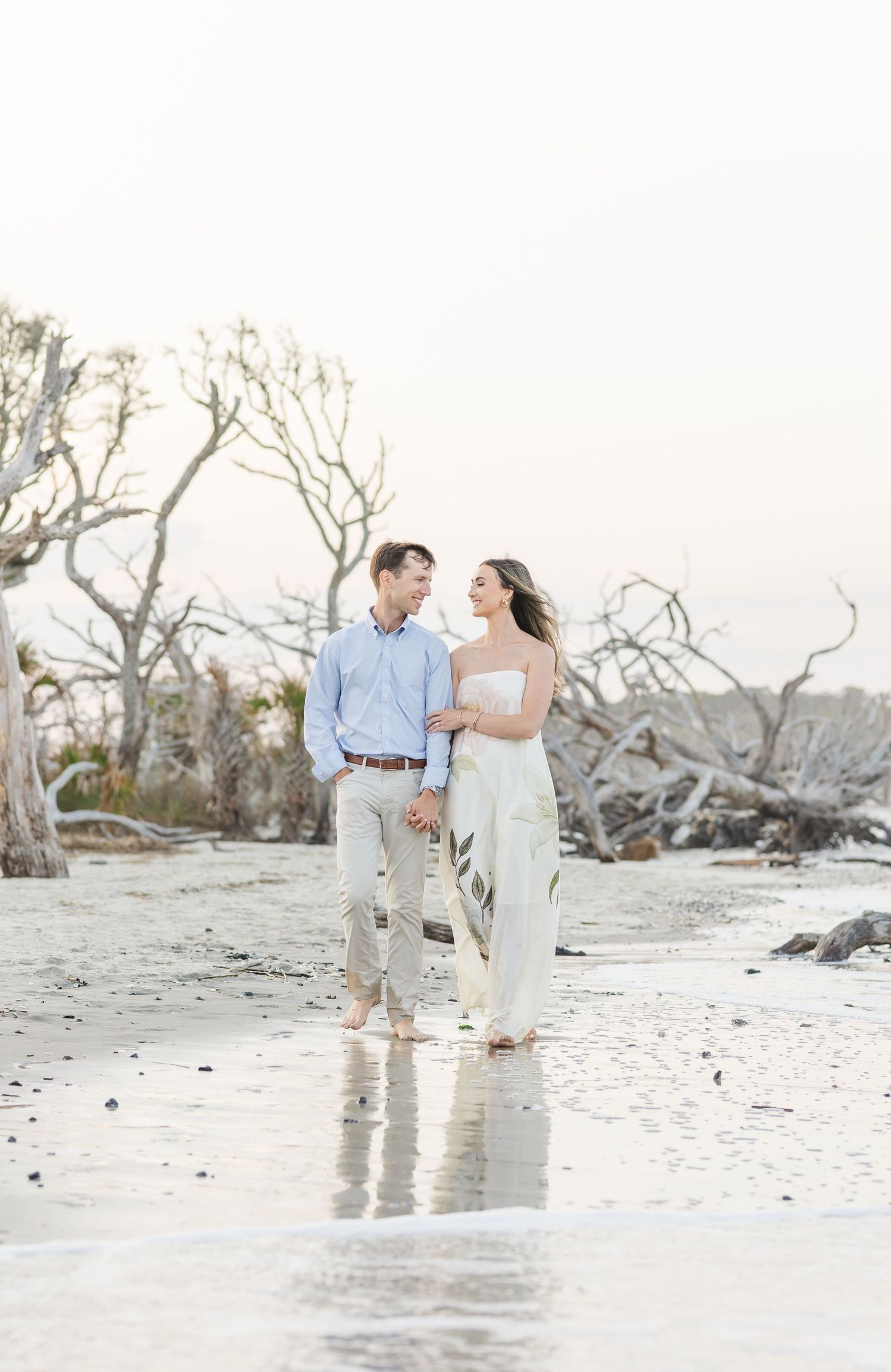 Couple holding hands walking barefoot on beach with driftwood trees in soft golden light.