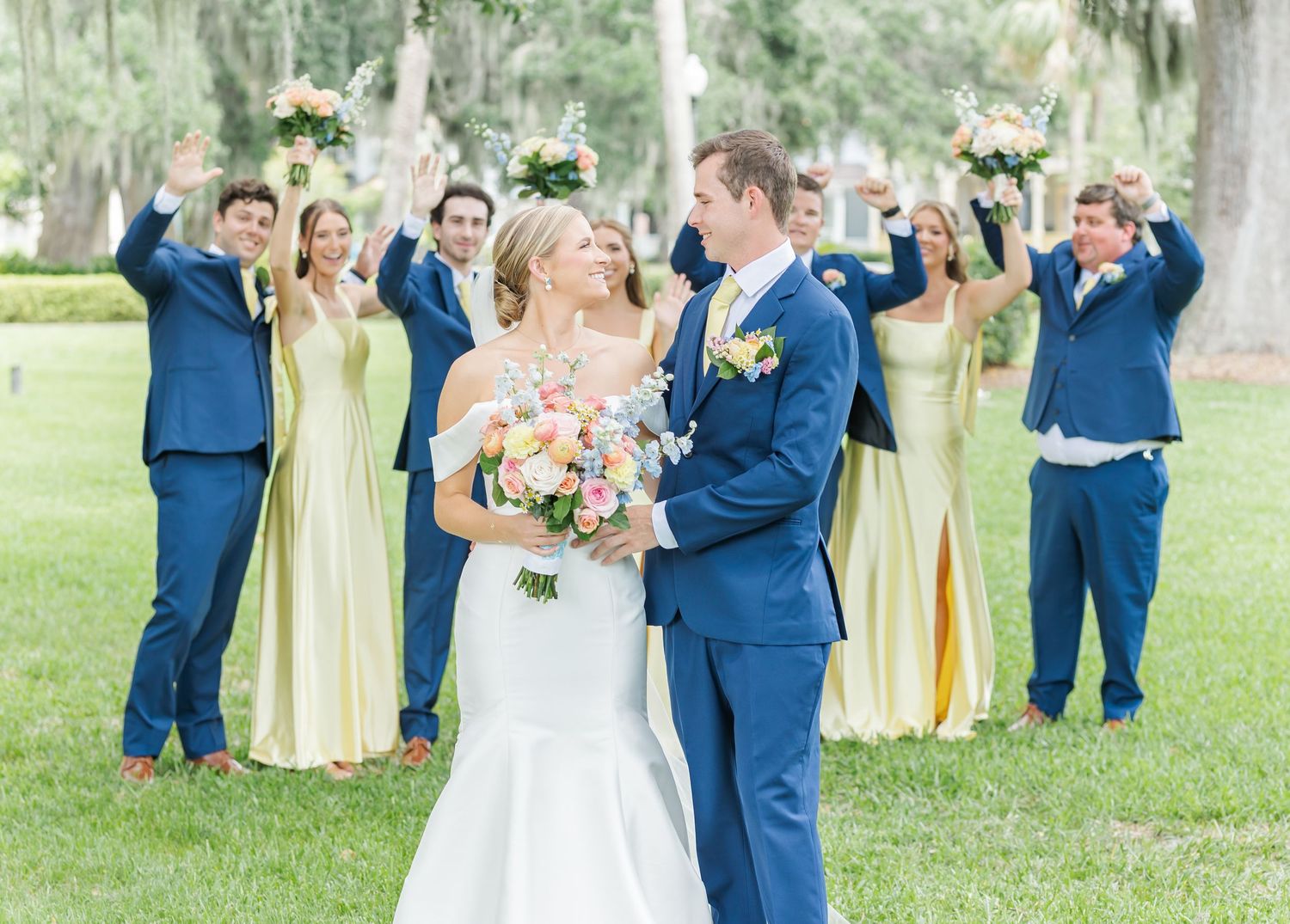 Wedding party in blue suits and yellow dresses celebrates on lawn with Spanish moss trees.