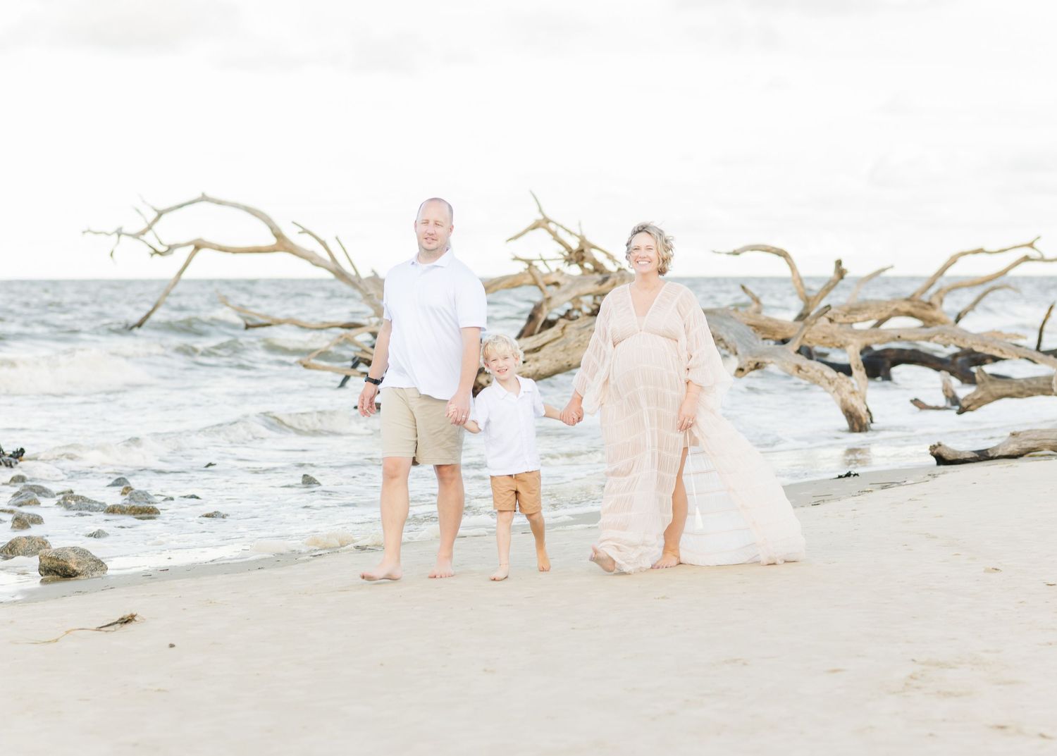 A couple walks along a scenic beach with driftwood and sand while wearing white clothing under cloudy skies.