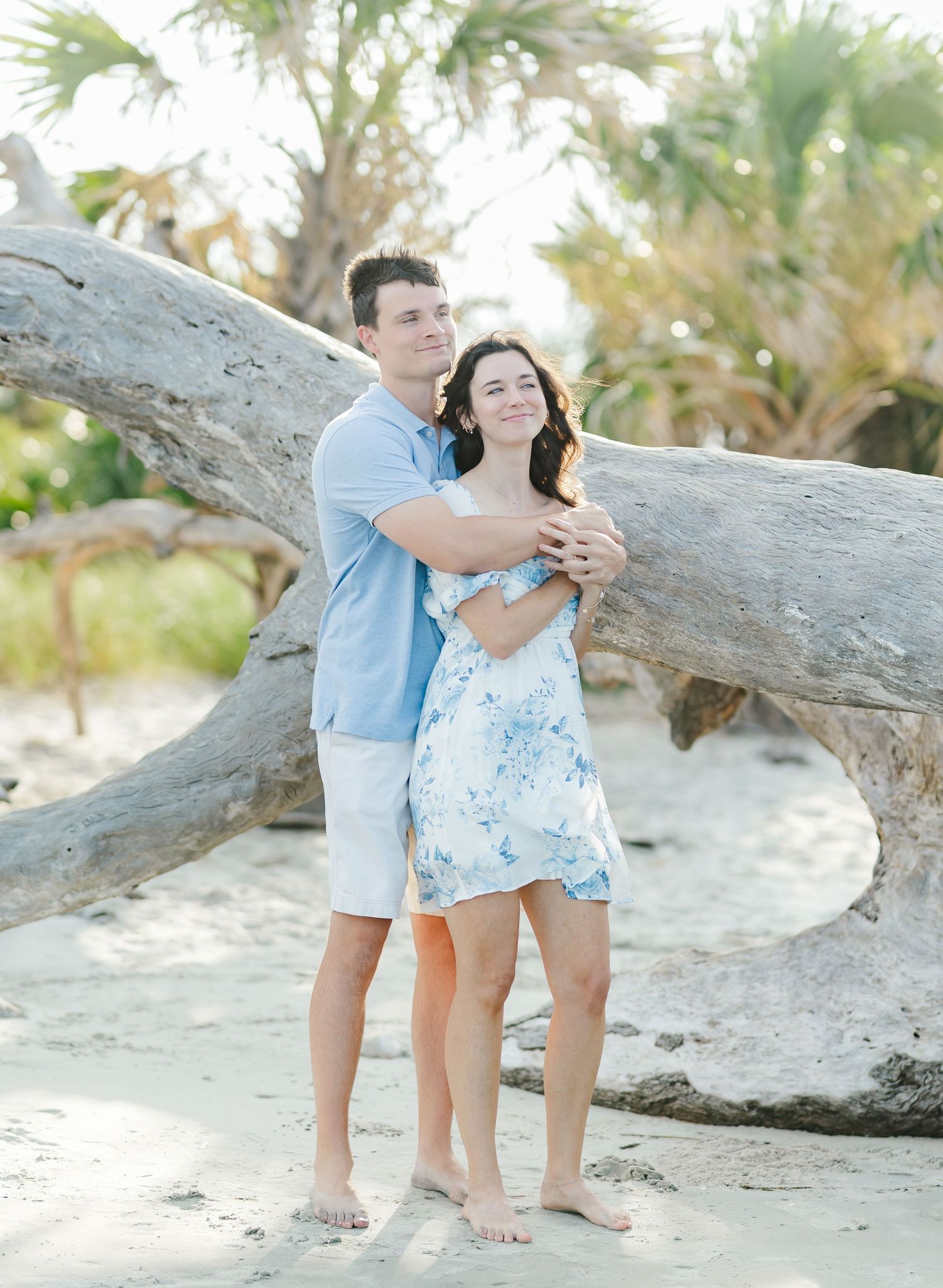 A couple shares a tender moment by a driftwood log on a sandy beach with palm trees.