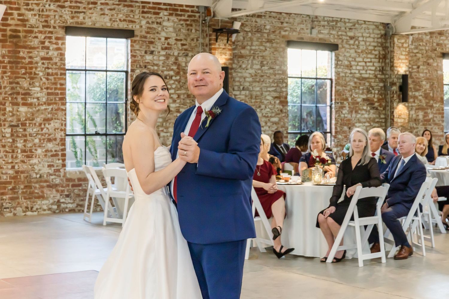 Wedding couple shares first dance in rustic brick venue with guests watching from white chairs.