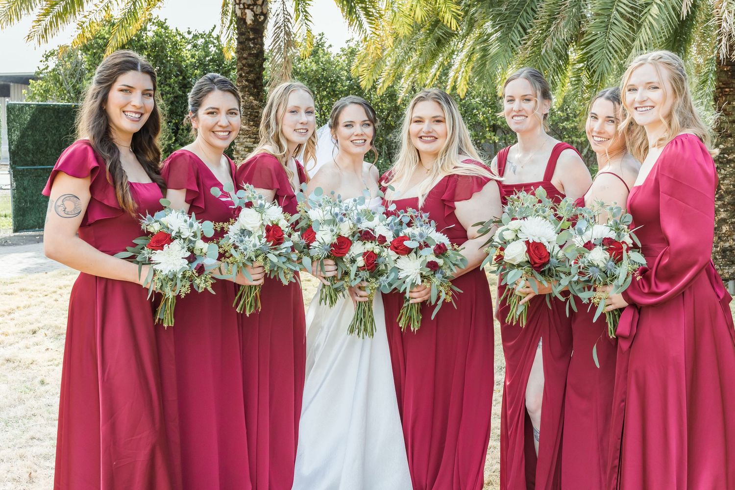 Wedding party in burgundy dresses holding red and white floral bouquets outdoors.