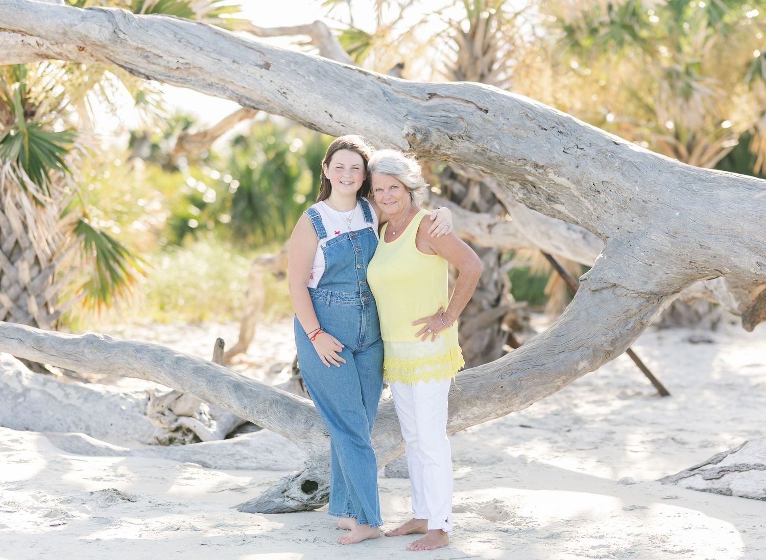 Two people stand leaning against a large driftwood log on a sunny beach with palm trees in the background.