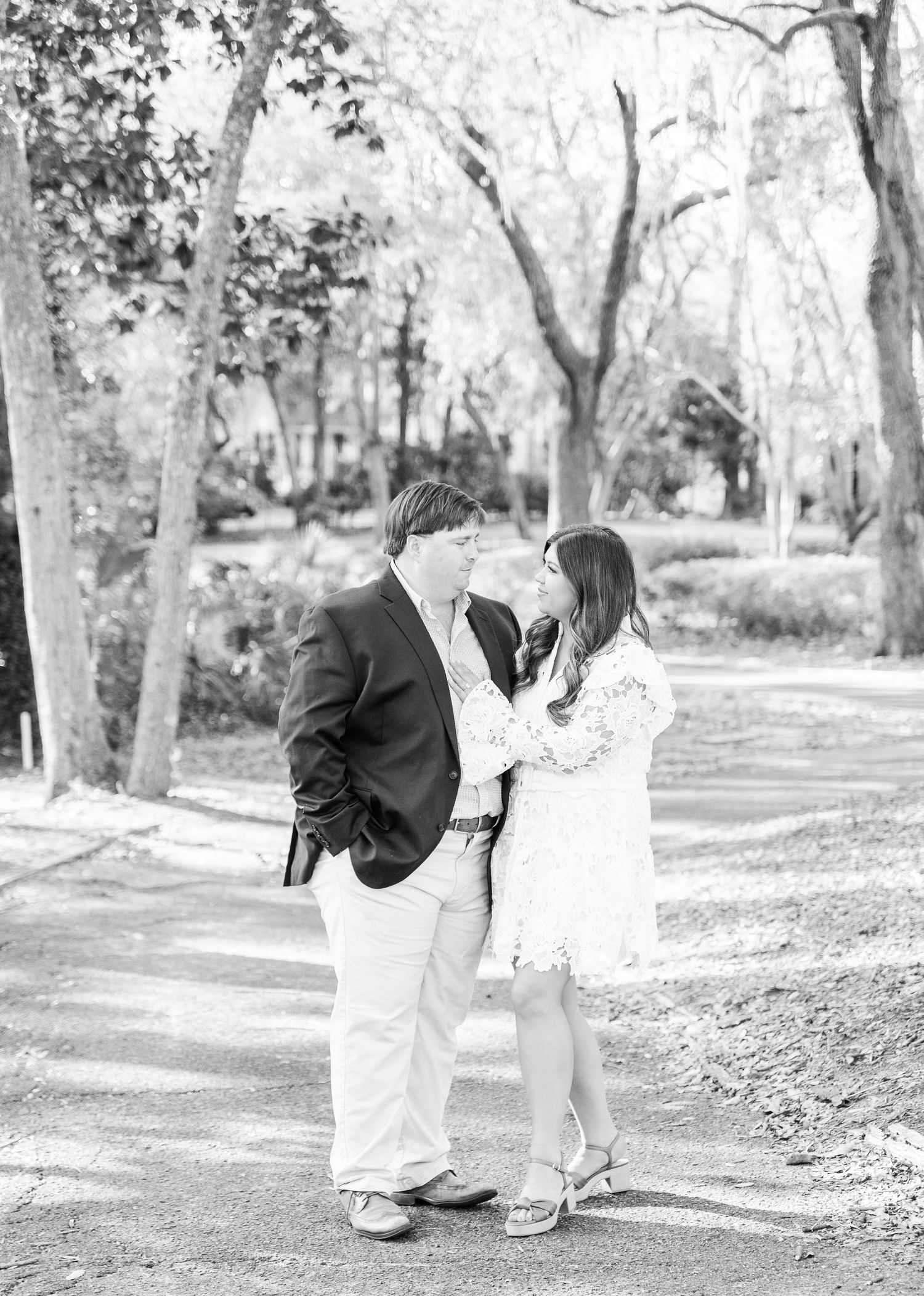 A romantic black and white engagement photo of a couple standing on a tree-lined path in a park setting.
