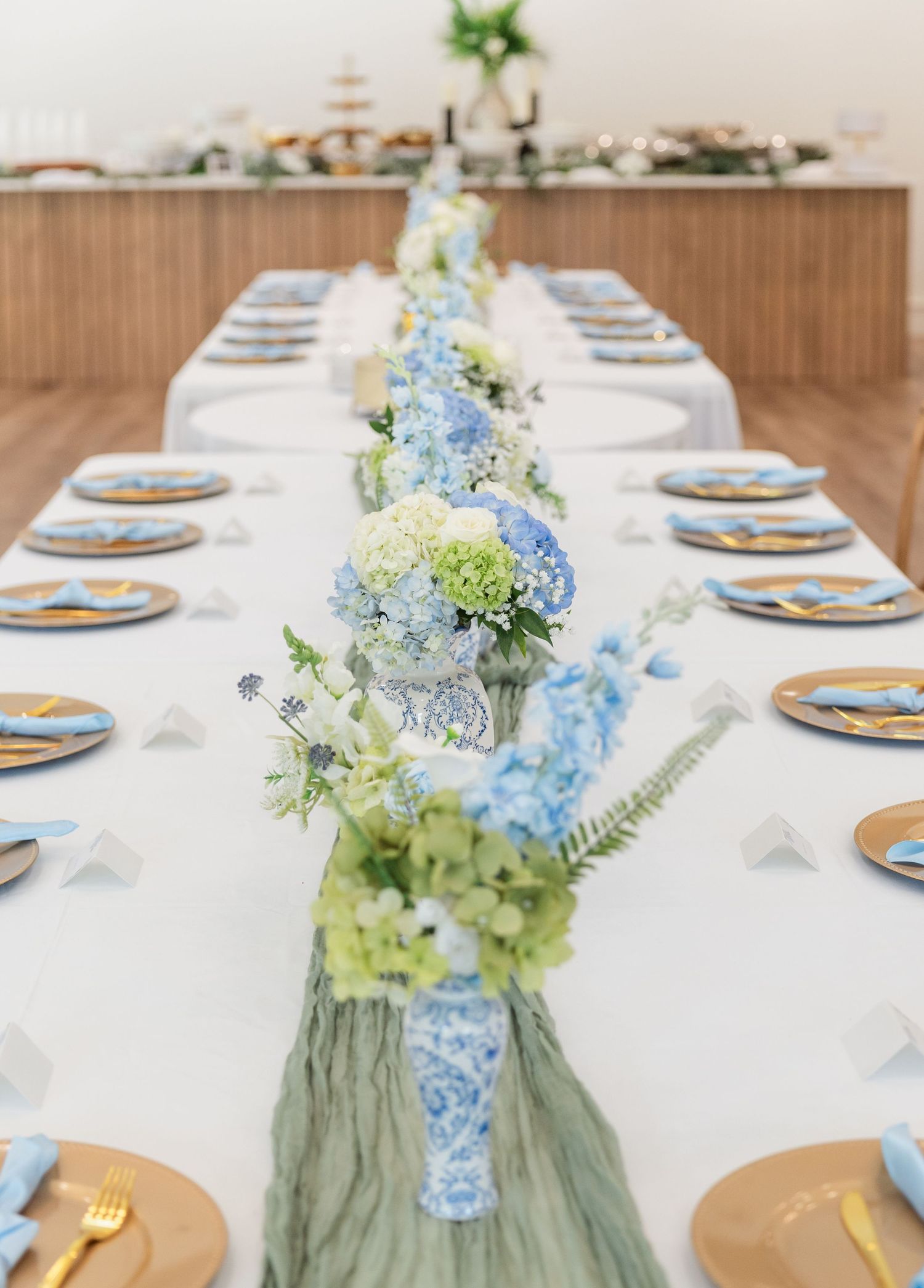 Long wedding reception table with blue and white hydrangea floral arrangements, gold chargers, and sage runner.