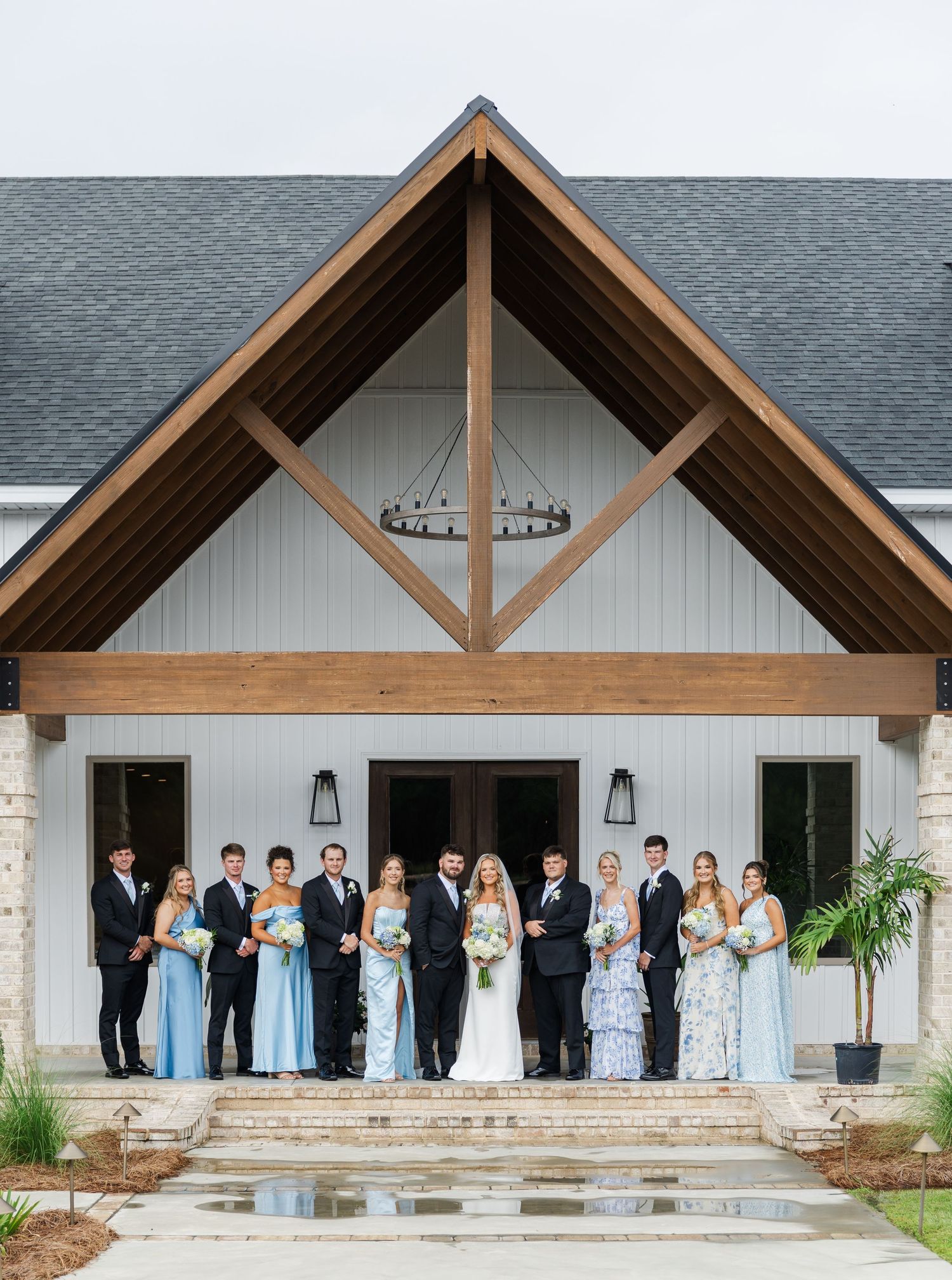 Wedding party in black suits and light blue dresses posing outside white barn chapel venue.