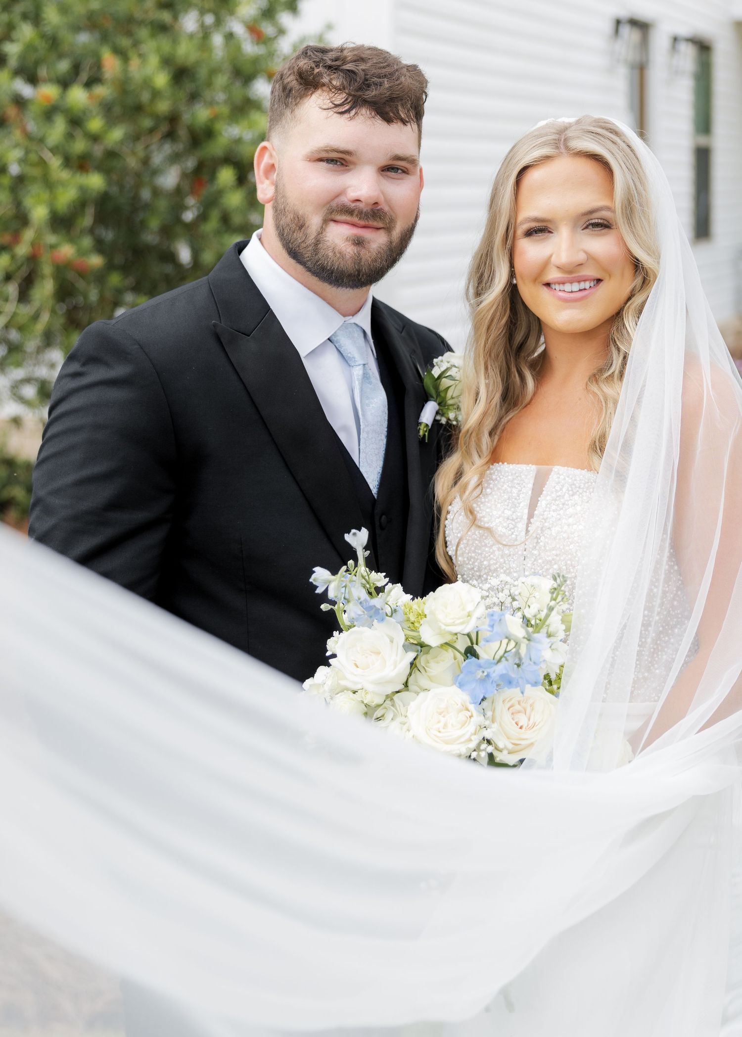 Smiling bride and groom posing outdoors, bride in pearl-embellished gown with veil flowing, holding white and blue bouquet.
