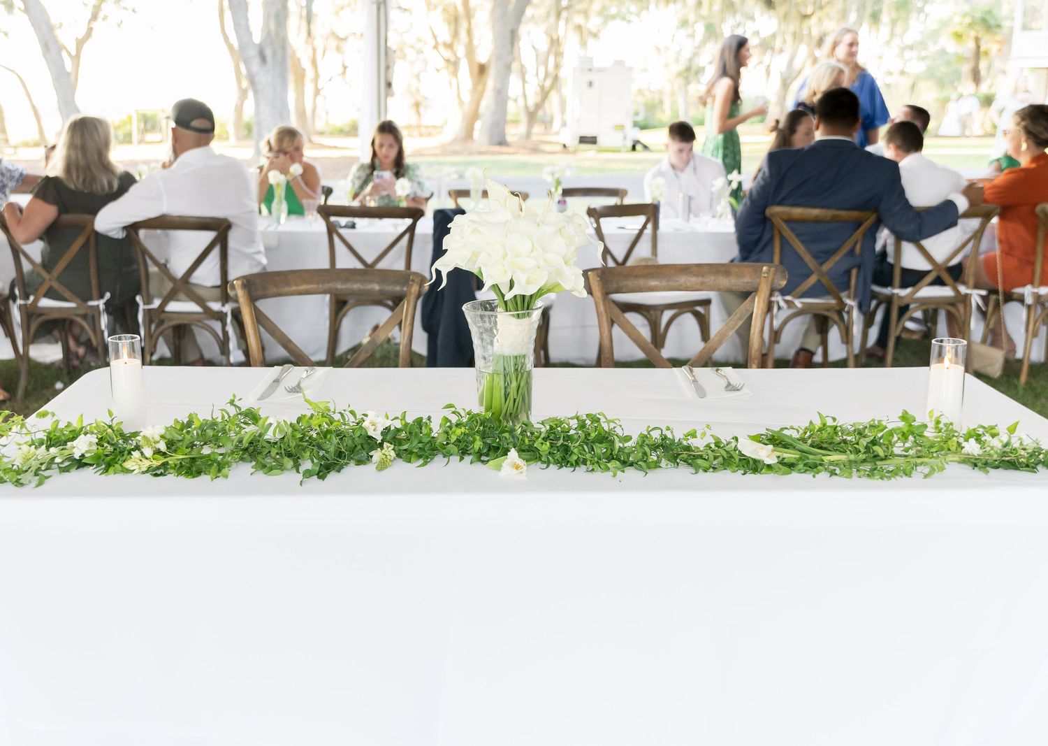 Wedding reception dining setup with cross-back chairs and white tablecloths under trees.
