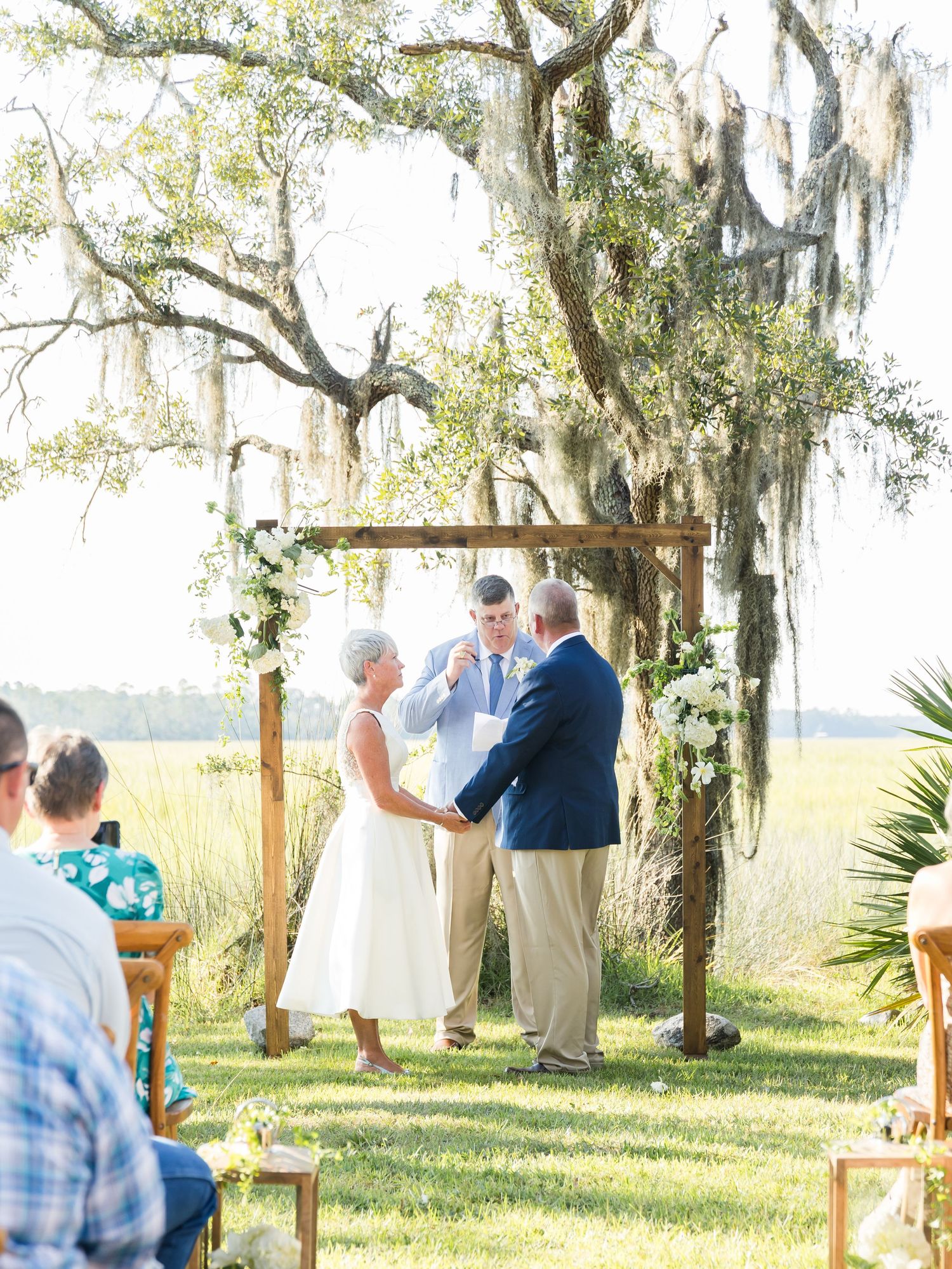 Outdoor wedding ceremony setup with wooden chairs and white fabric decorations under oak trees.