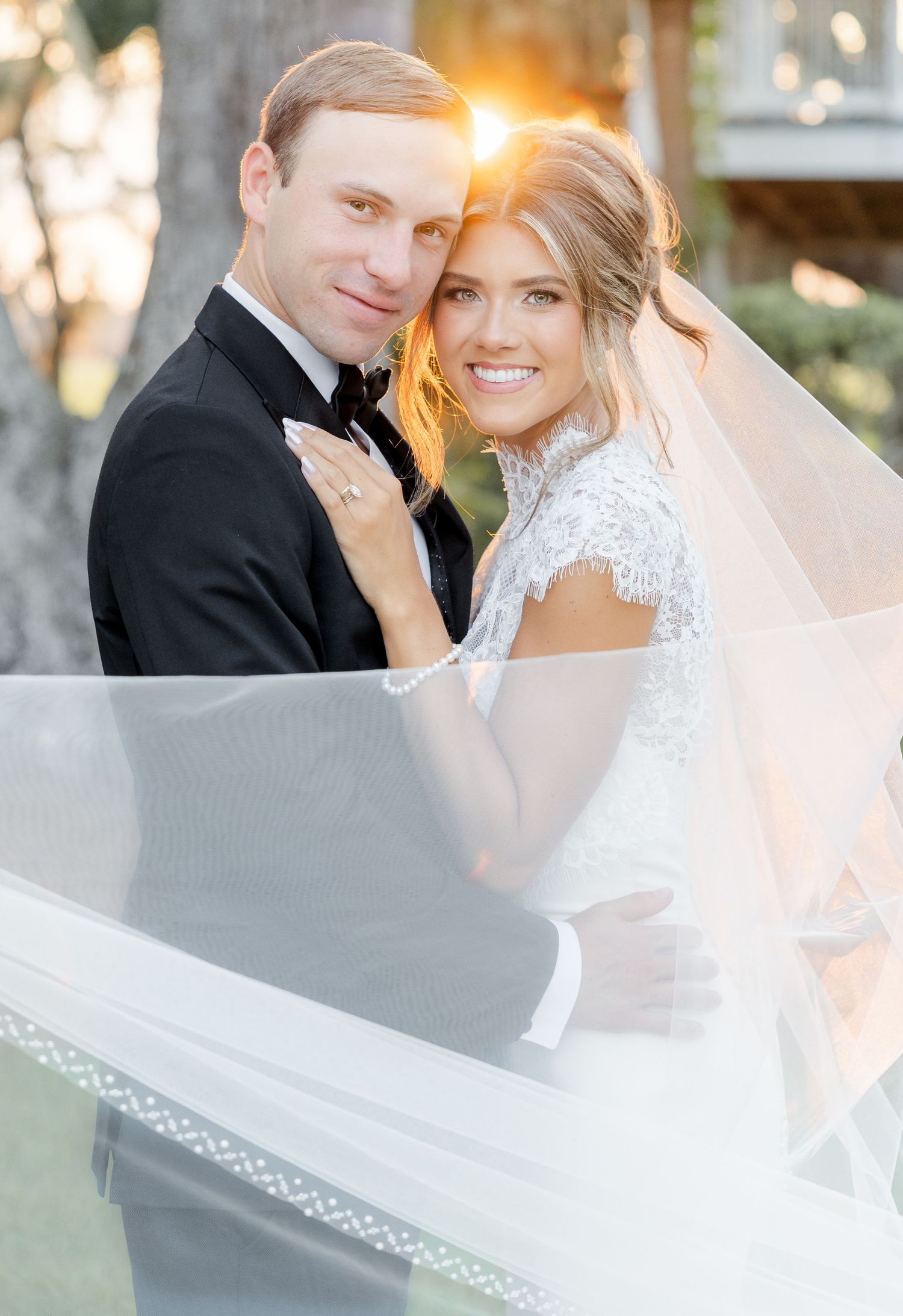 Bride and groom pose together outdoors at golden hour, bride wearing lace dress and flowing veil, groom in black tuxedo.
